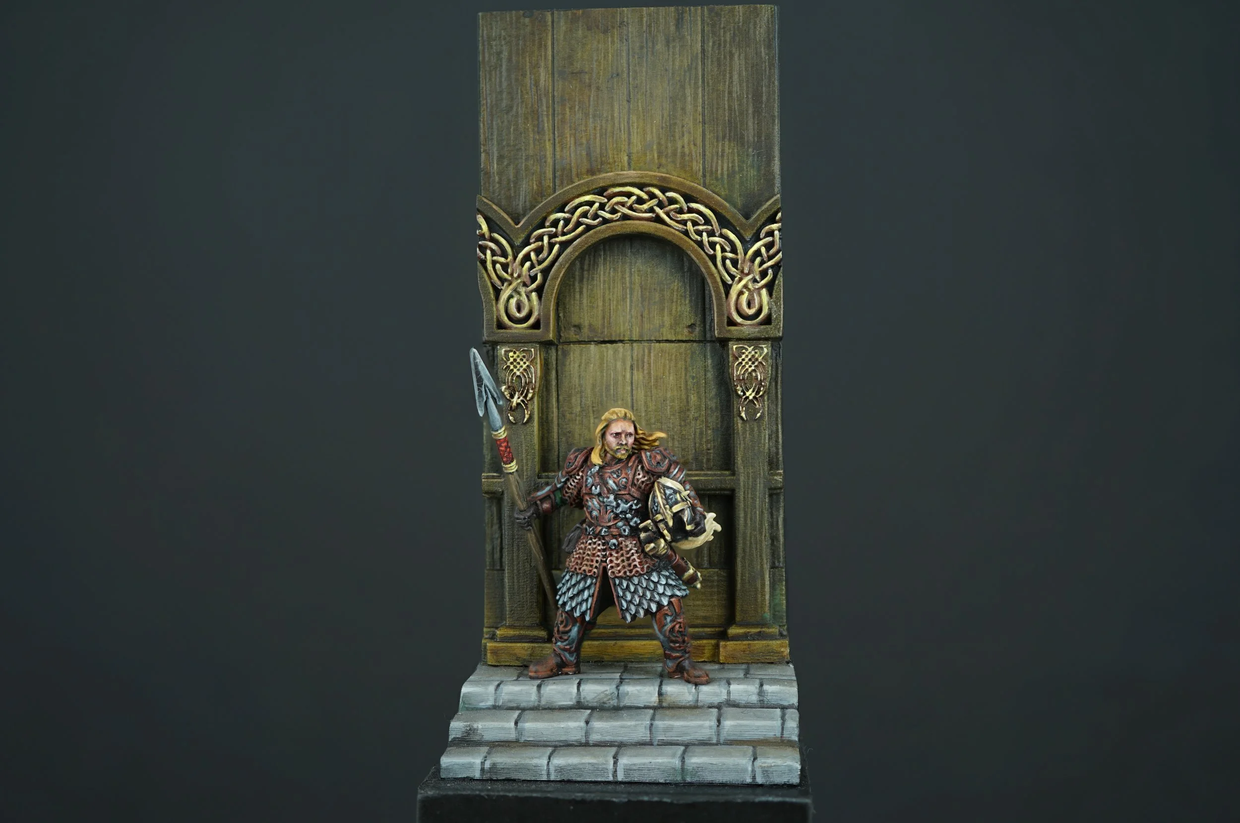 Miniature figure of Eomer from The Lord of The Rings standing on stone steps in front of a large wooden door representing the golden hall of Meduseld with ornate carvings.