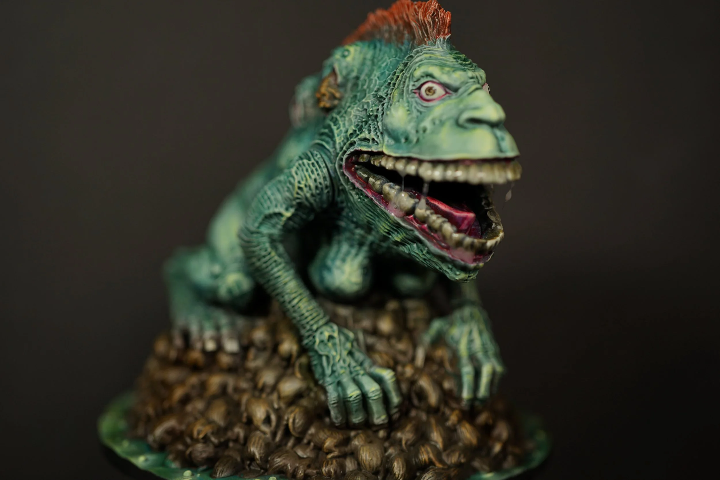 A detailed figurine of a FrogDog from the Kingdom Death franchise with a human face, open-mouthed revealing sharp teeth, seated on a rocky base.