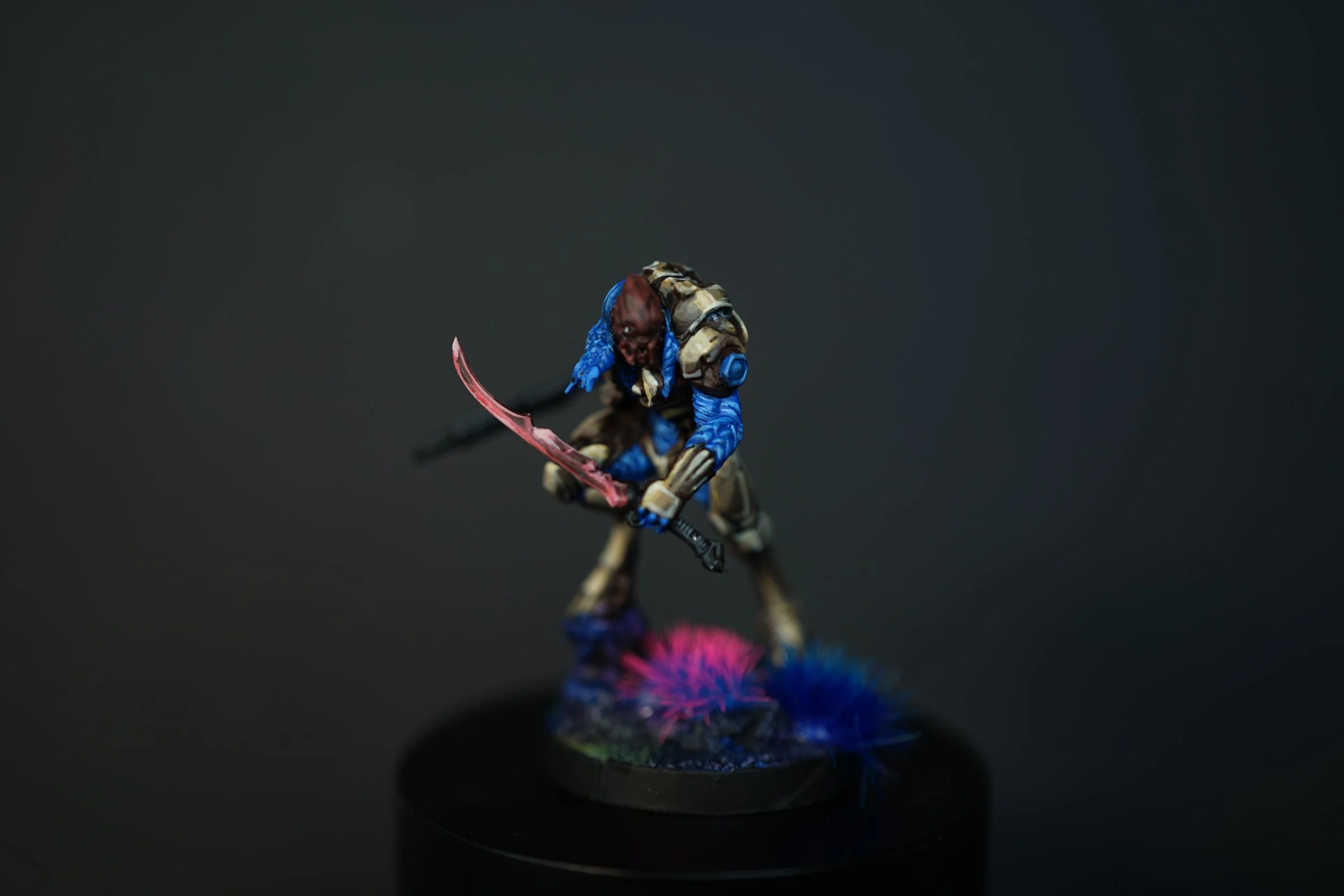 Miniature figurine of a fierce alien creature from the Infinity game with a bald head, blue and tan armor, wielding a pink-bladed weapon, standing on a circular base decorated with pink and blue plants against a dark background.