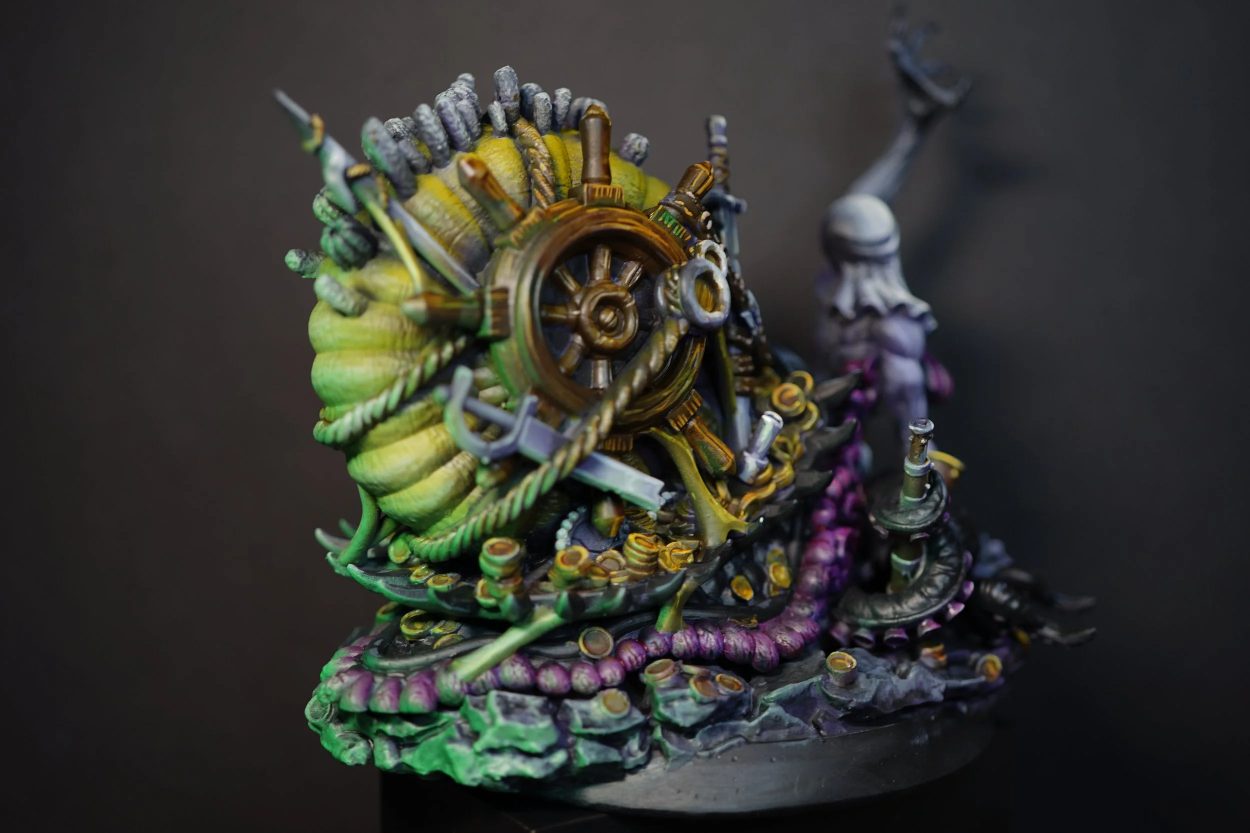 A detailed sculpture featuring a coiled sea creature with metallic and organic elements, including a wheel, ropes, and tentacles, set against a dark background.