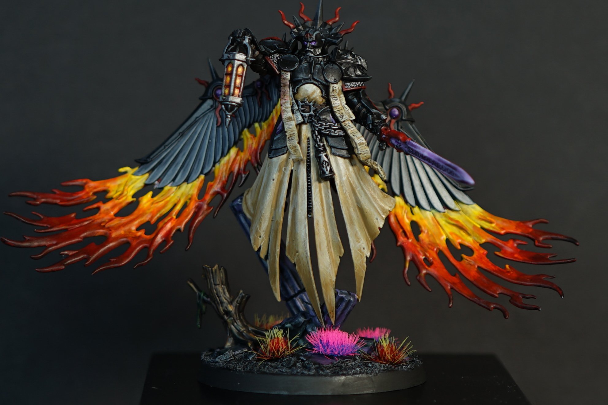 A detailed miniature figurine of a dark angel with large fiery wings, wearing armor and a flowing robe, holding a sword and lantern, mounted on a decorated base with pink and red plant-like elements.
