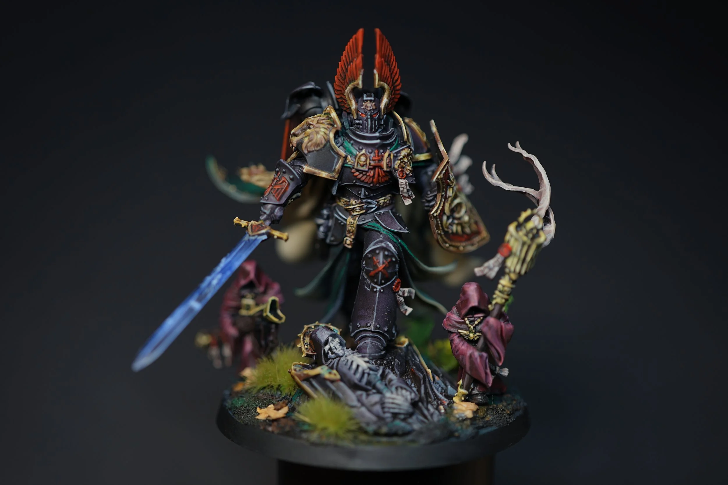 Detailed miniature figure of a dark armored warrior with a red crest, holding a blue sword, standing over a skeletal corpse, surrounded by robed figures, set on a textured base with grass and leaves against a black background.
