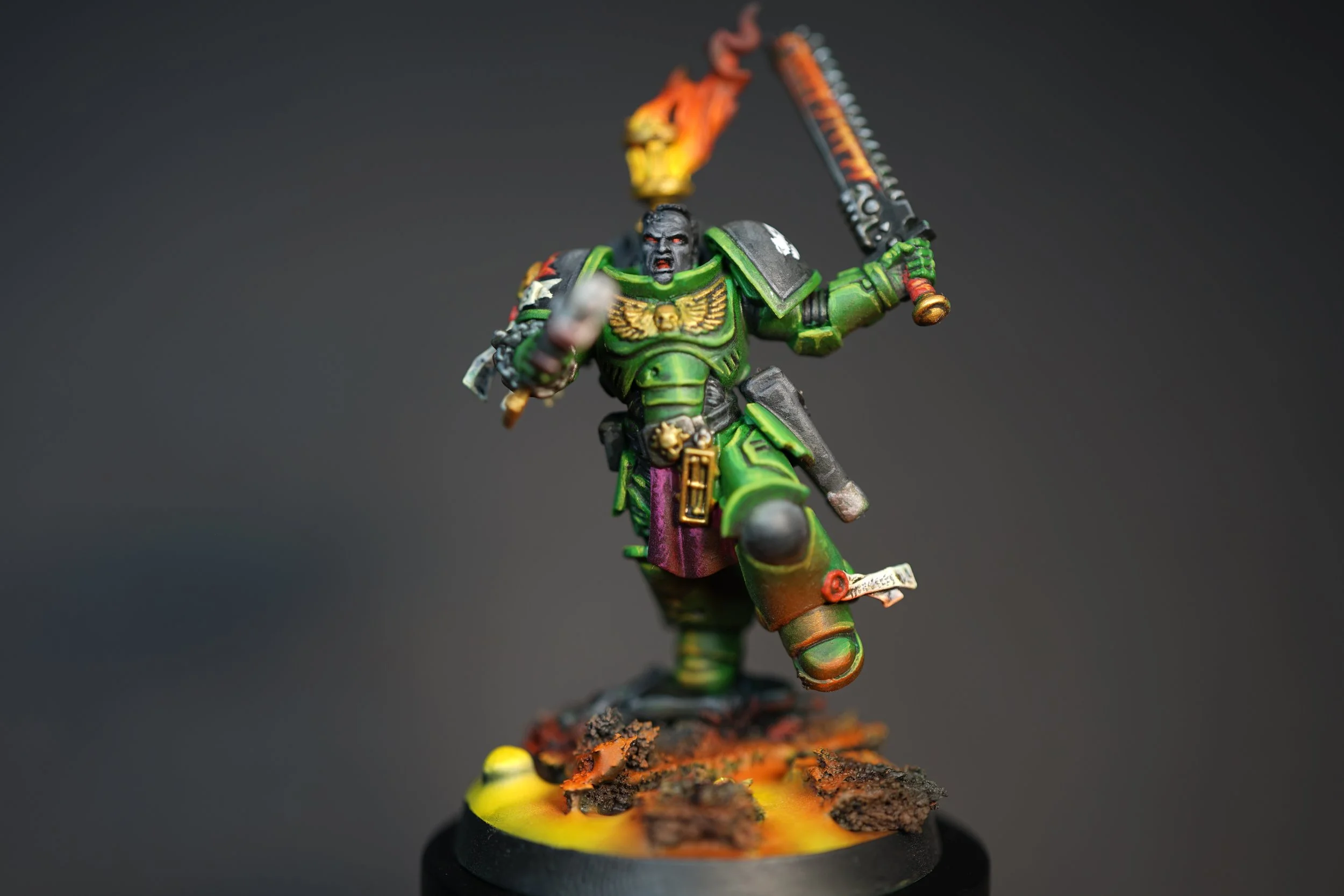 Miniature figurine of lieutenant Titus from Warhammer 40k in green armor wielding a flaming chainsword and a gun, standing on a decorated base with debris and lava effects.