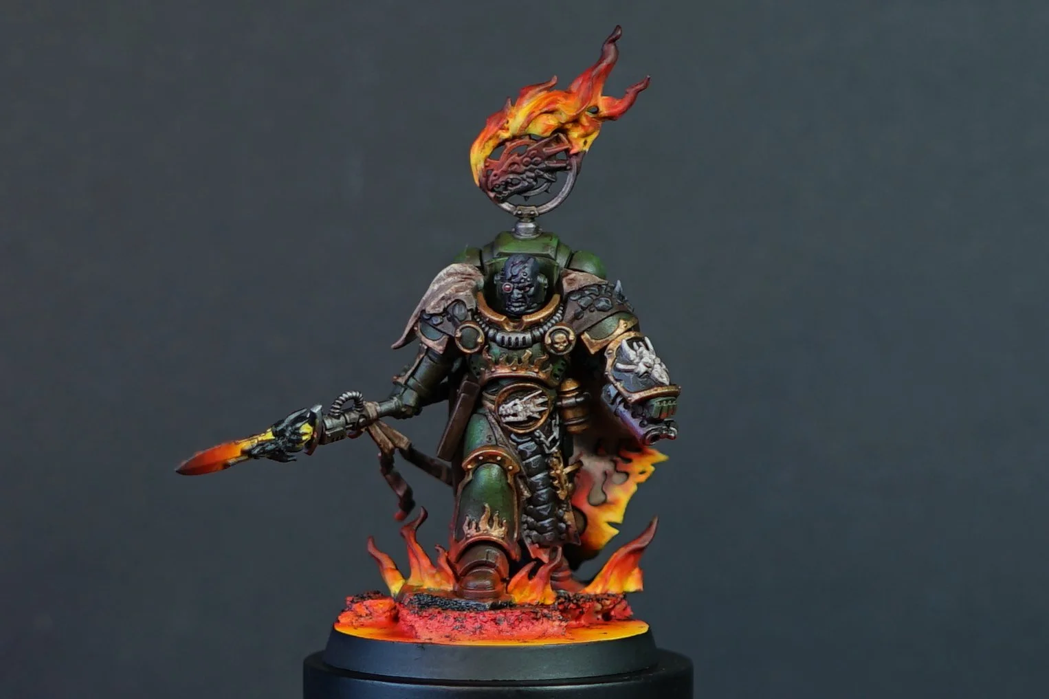 A painted miniature figure of Vulkan He'Stan from Warhammer 40k in dark green armor, holding a flaming spear, standing on a base with fire effects, with a large flaming fireball above his head, set against a plain dark background.