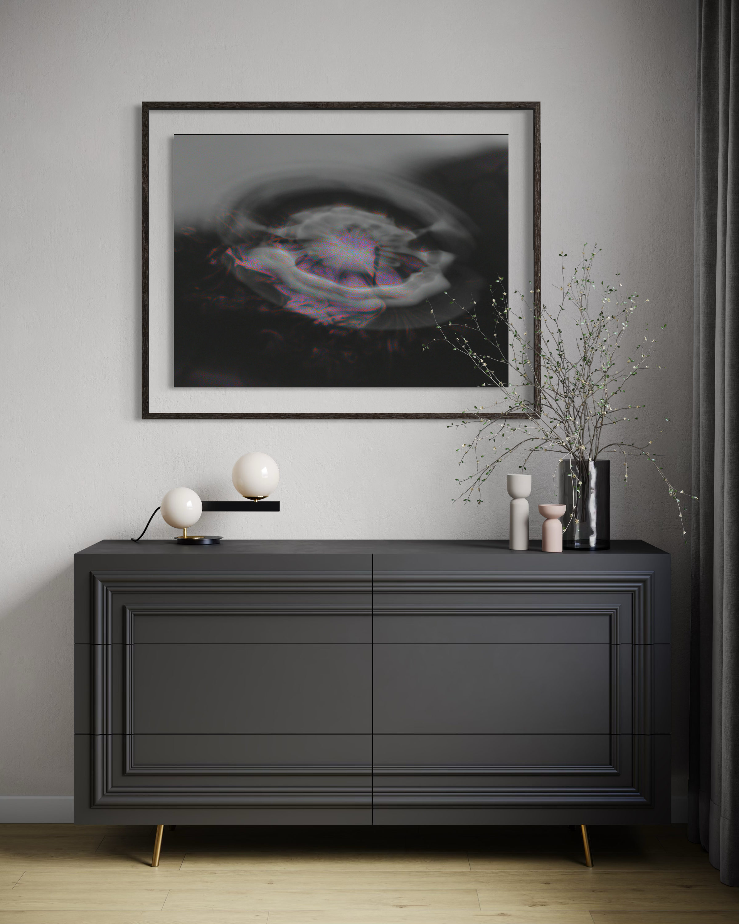 A modern gray sideboard or cabinet with decorative objects on top, including two white spherical lamps, a black vase with twigs, and a white and pink vase. Above the sideboard is a large framed black and white abstract artwork featuring swirling patterns and a splash of color.