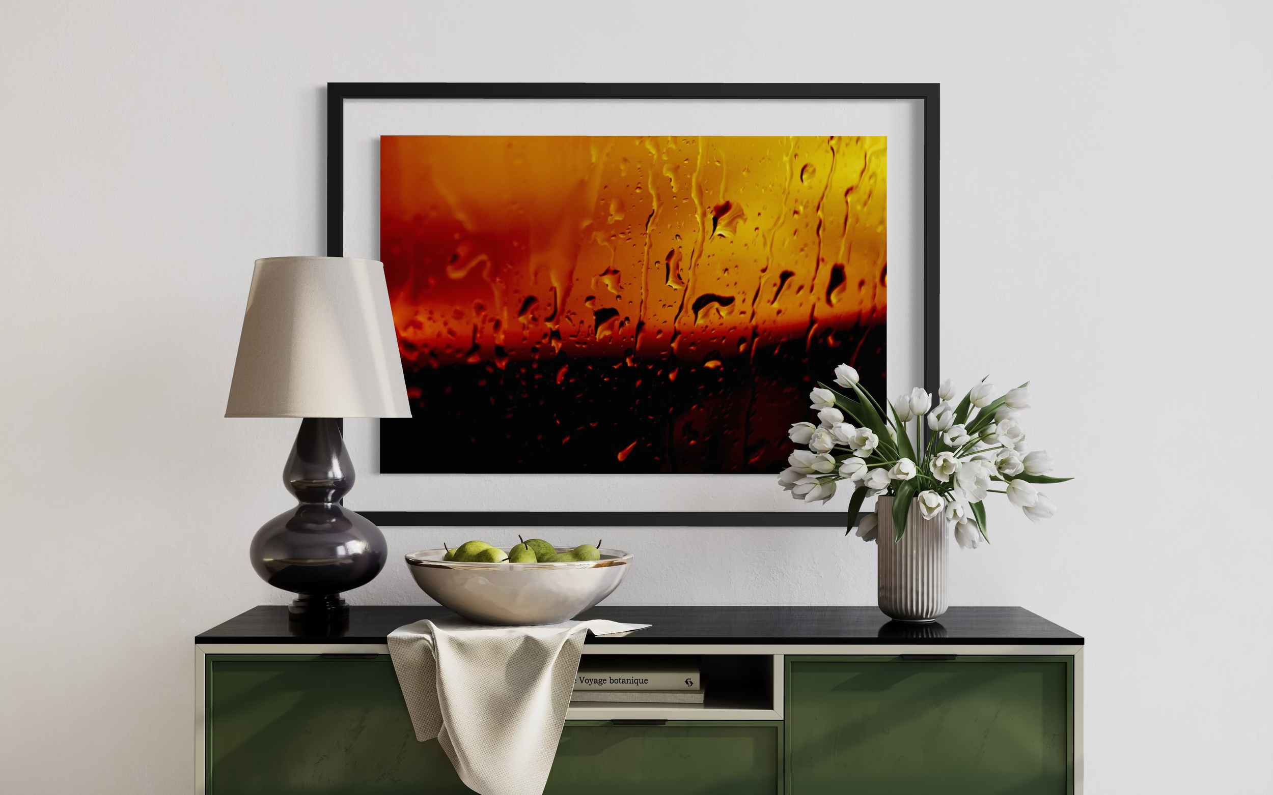 Living room with black framed artwork featuring abstract hot air balloon scene, white flowers in a striped vase, green apples in a bowl, white lamp, and a green and black cabinet.