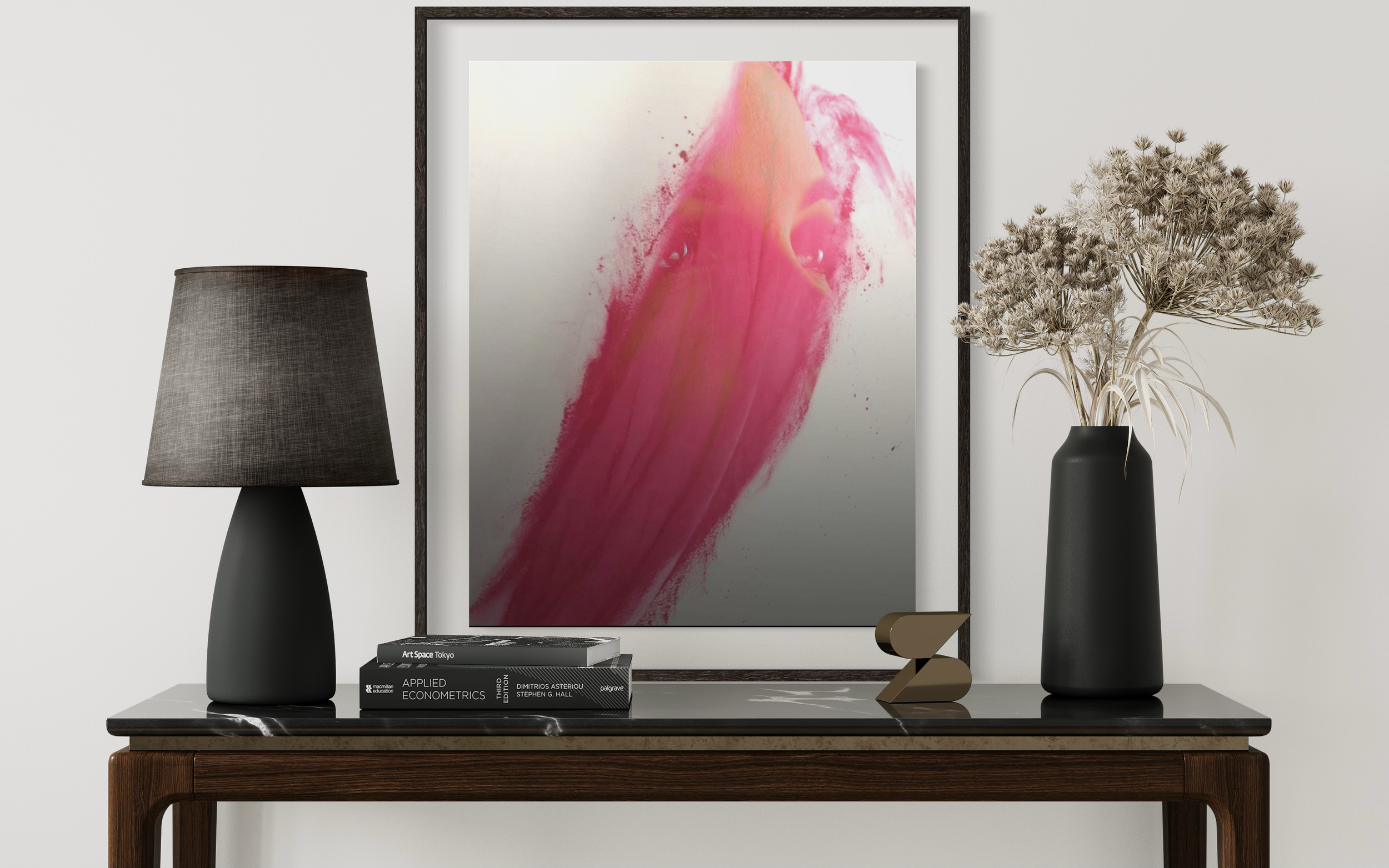 A modern interior decor setup featuring a framed abstract pink and red artwork, two black vases with dried flowers, a grey textured table lamp, a small gold decorative object, and a stack of black books on a wooden table.