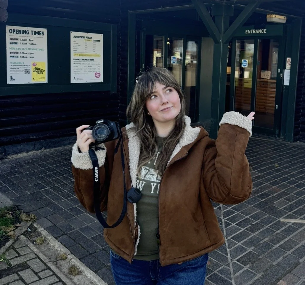 A young woman with shoulder-length brown hair wearing a brown shearling-lined jacket, holding a Canon camera in her right hand, standing in front of an entrance to a building with signs displaying opening times and admission prices.