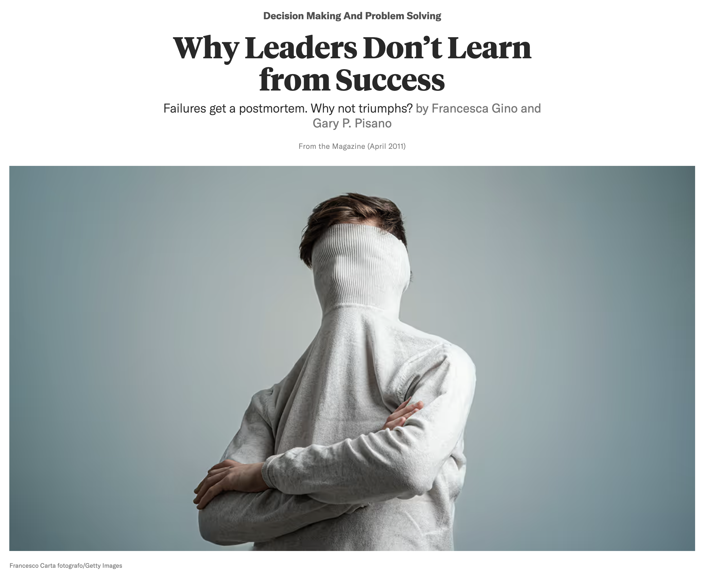 A person with short brown hair wearing a gray sweatshirt and covering their face with the sweatshirt, standing with arms crossed against a plain muted background.