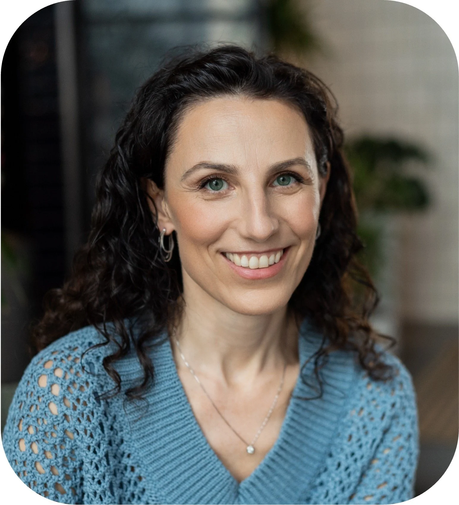 A woman with curly dark hair and blue eyes, smiling, wearing a blue knitted sweater and a silver necklace, indoors with blurred background.