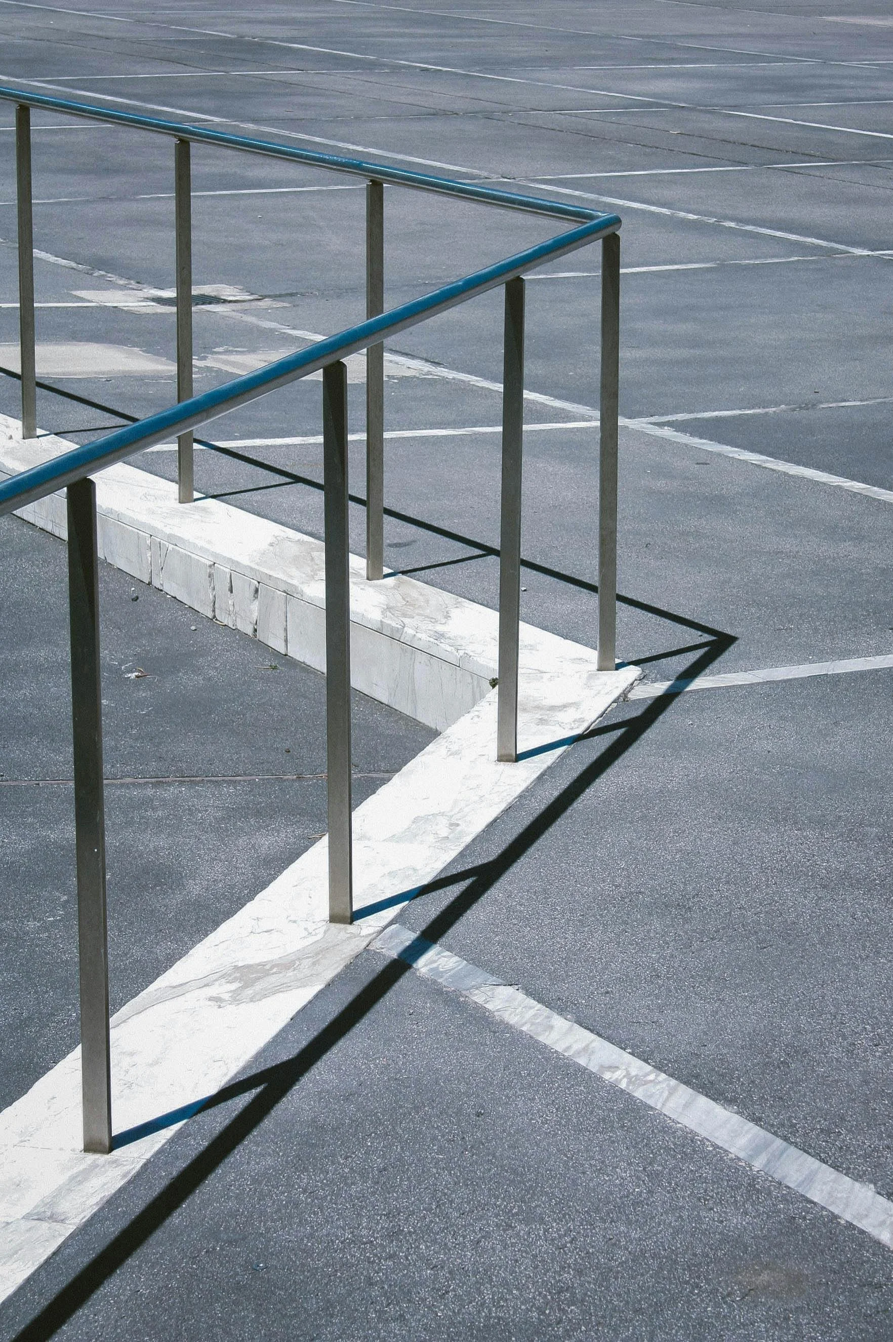 An empty parking lot with marked spaces and a metal railing casting a shadow on the asphalt.