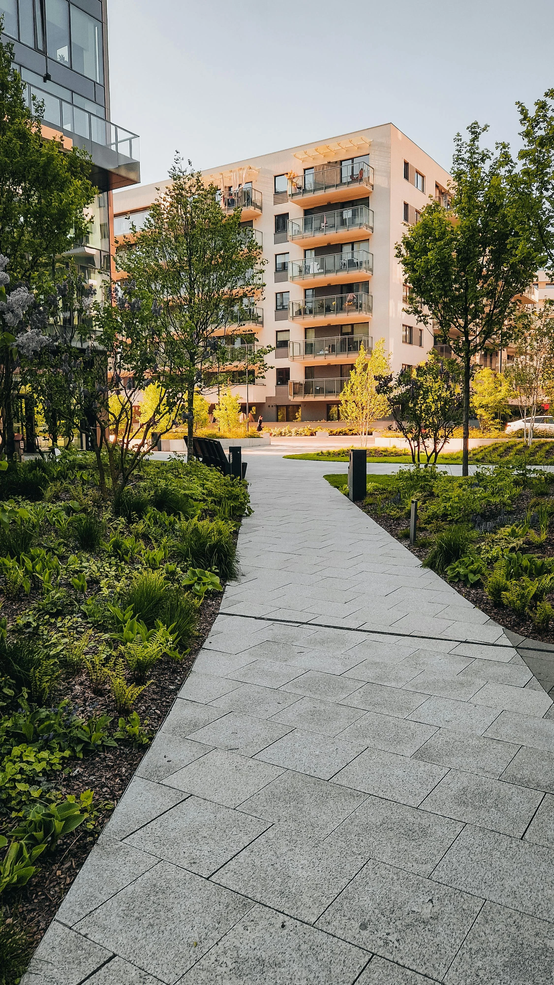 A concrete pathway winds through a landscaped green space with trees and shrubs, leading to a modern apartment building with balconies and large windows, under a clear sky.