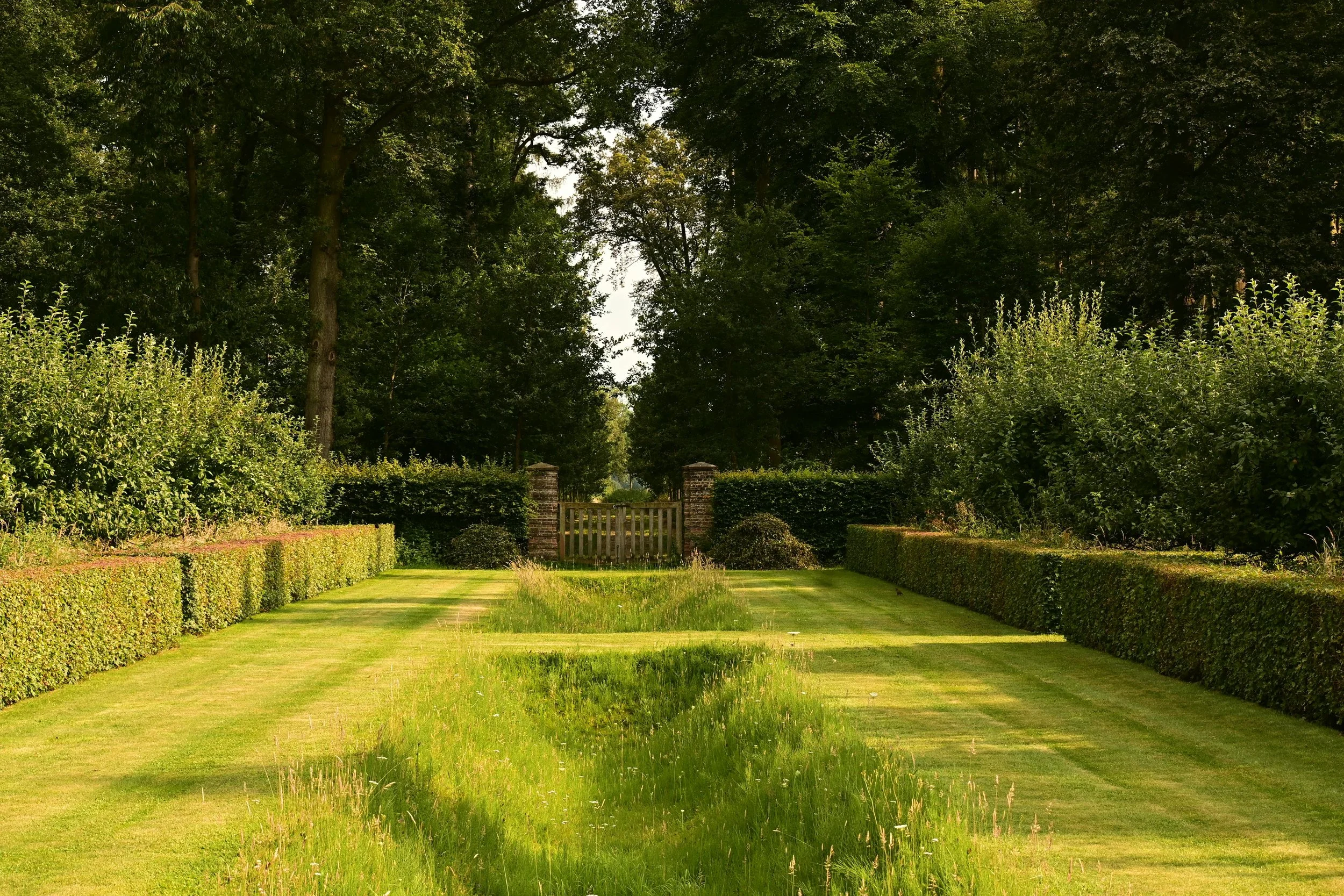 A well-maintained garden with a grassy path, trimmed bushes on each side, and a wooden gate in the distance, surrounded by tall green trees.
