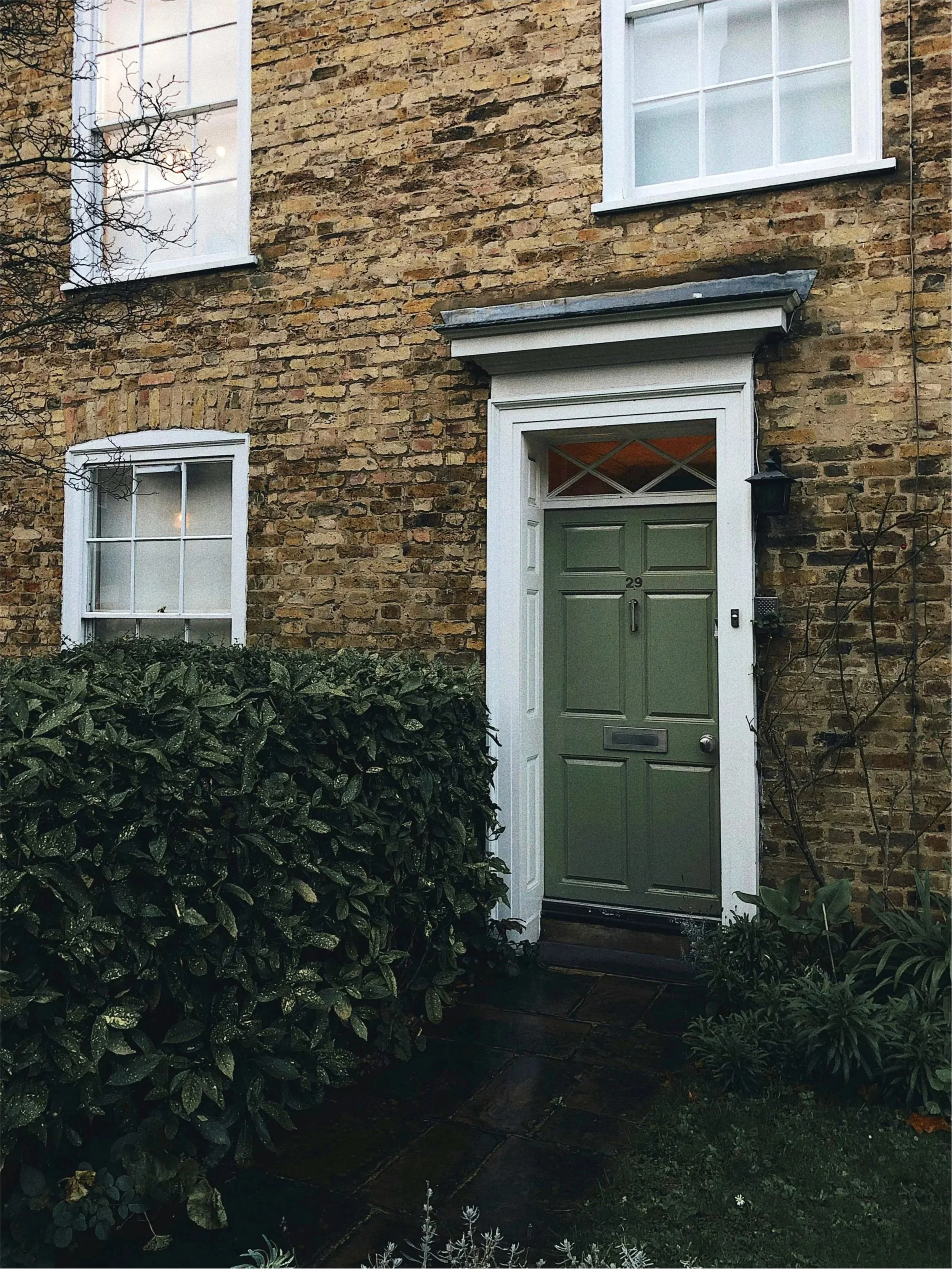 A brick house with two white-framed windows on the upper floor and one on the ground level, with a green front door numbered 29. There are bushes and plants in front of the house, and a small tree to the left side of the image.