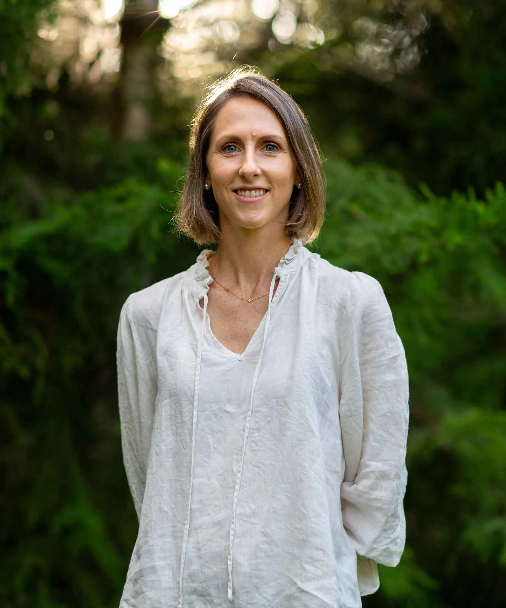 A woman with light brown hair and blue eyes smiling with trees behind her.