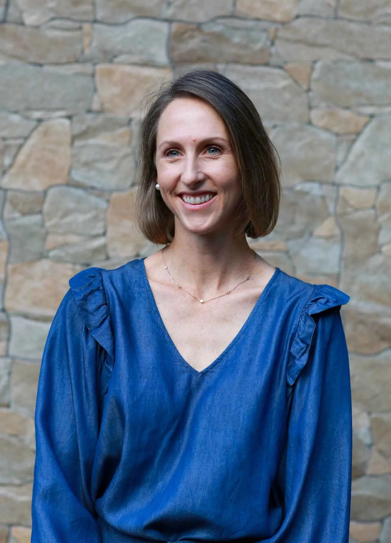 A woman with short brown hair and blue eyes, standing in front of a stone wall.