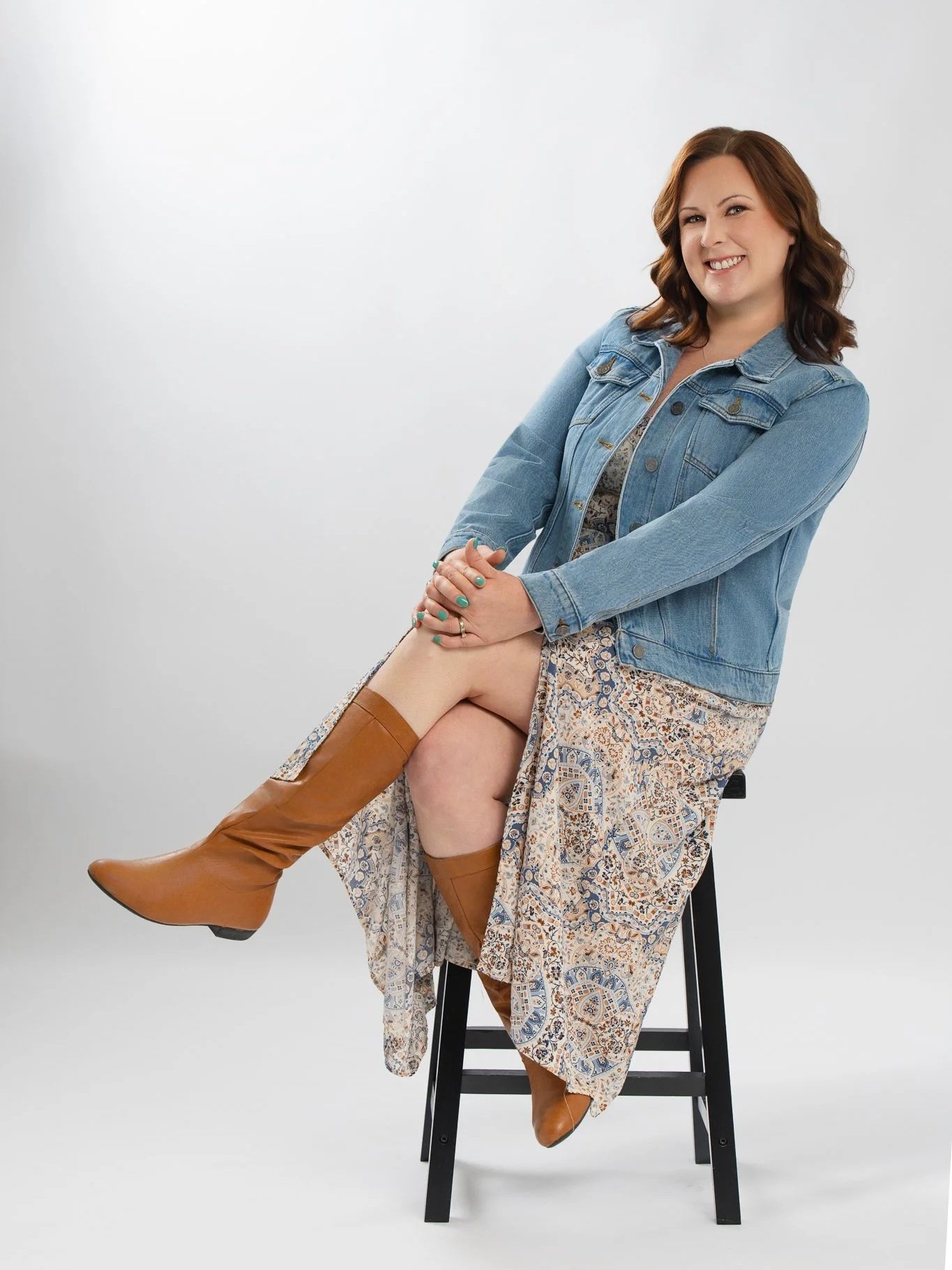 A woman sitting on a black stool, smiling, with her nude leg elevated and bent at the knee, wearing a floral dress, a denim jacket, and tall brown boots, against a plain white background.