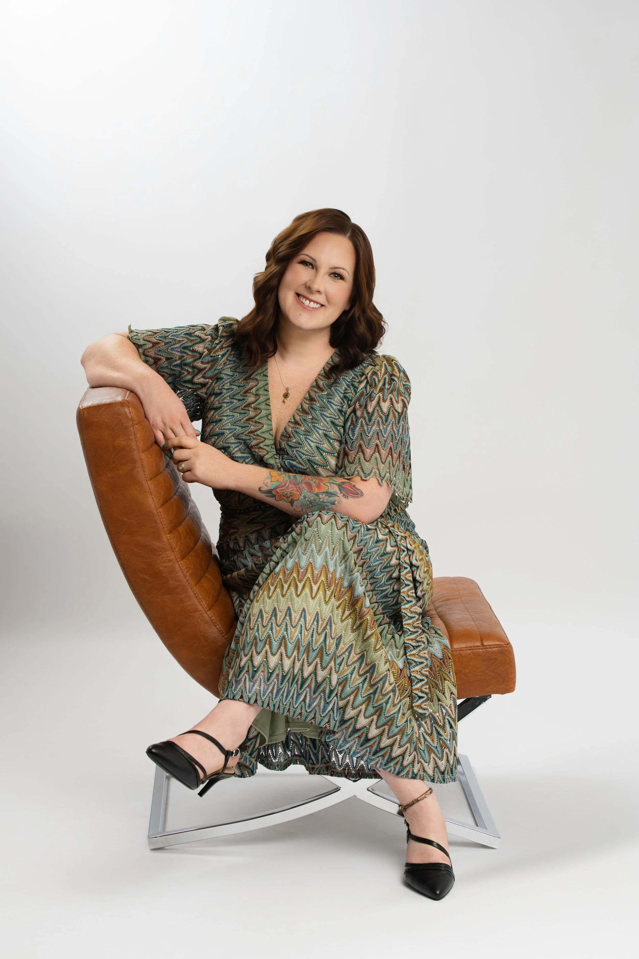 A woman sitting on a modern brown leather chair with metal legs against a plain white background. She has shoulder-length wavy brown hair, is smiling, and is wearing a colorful patterned dress with short sleeves and a V-neck. She has a tattoo on her left forearm, a necklace, and black high-heeled shoes with ankle straps.