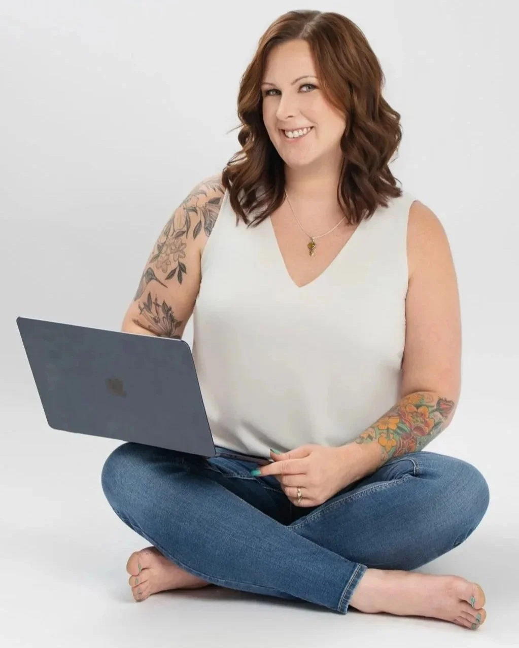 A smiling woman with shoulder-length wavy brown hair, tattoos on her arms, wearing a white sleeveless top and blue jeans, sitting cross-legged on the floor holding a laptop in front of her against a plain white background.