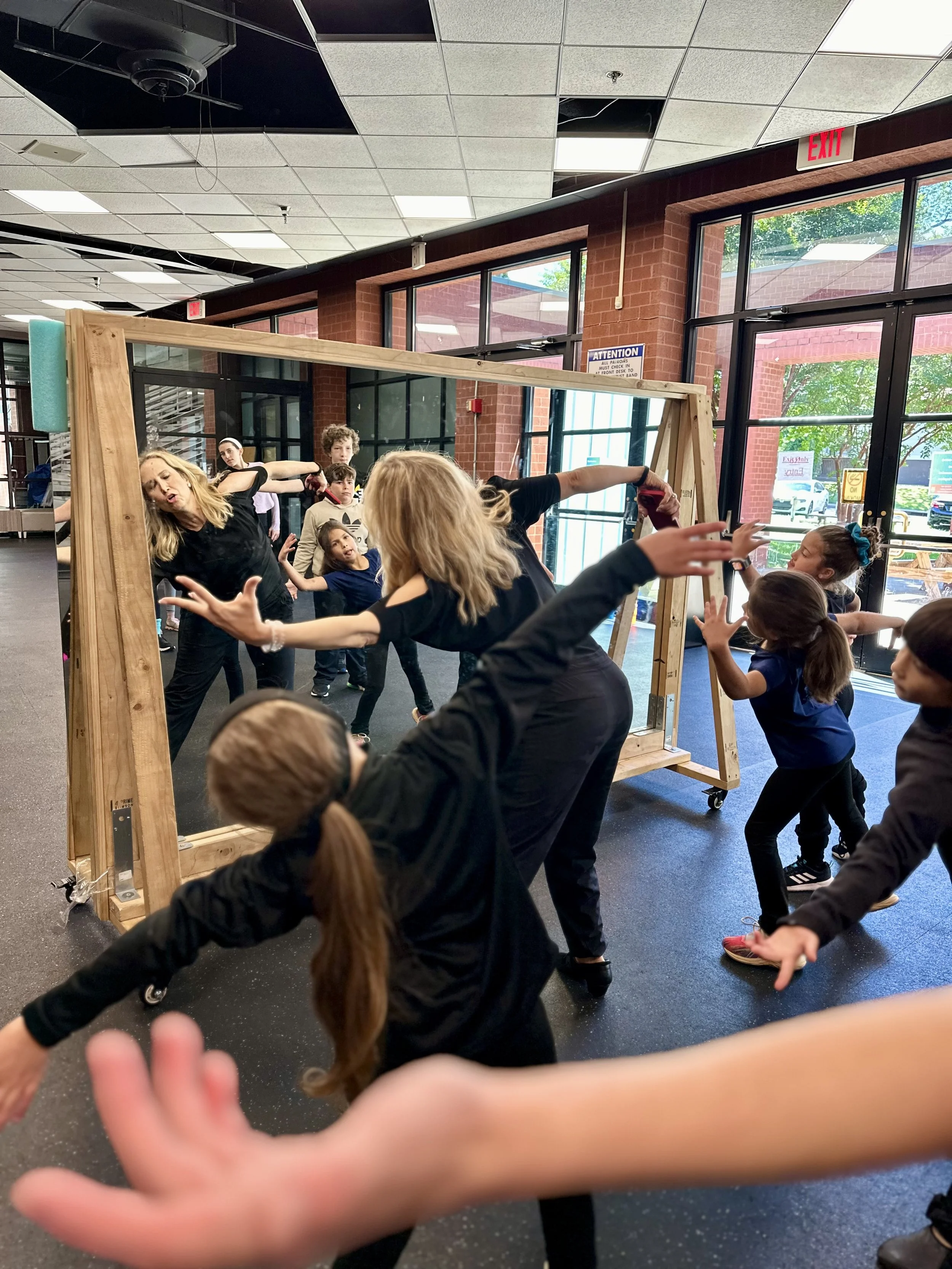 A group of children and a woman are playing with a large wooden mirror frame indoors, reaching out and making faces.