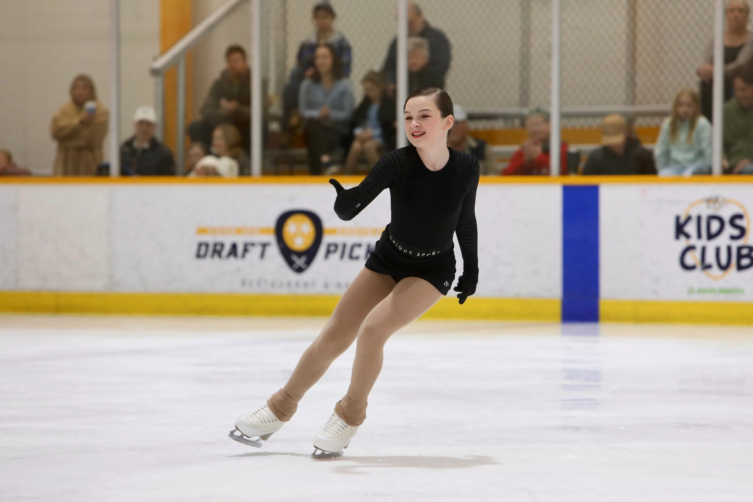 A young female figure skater in black attire with beige tights and white skates performs on ice, with an audience watching behind protective glass.