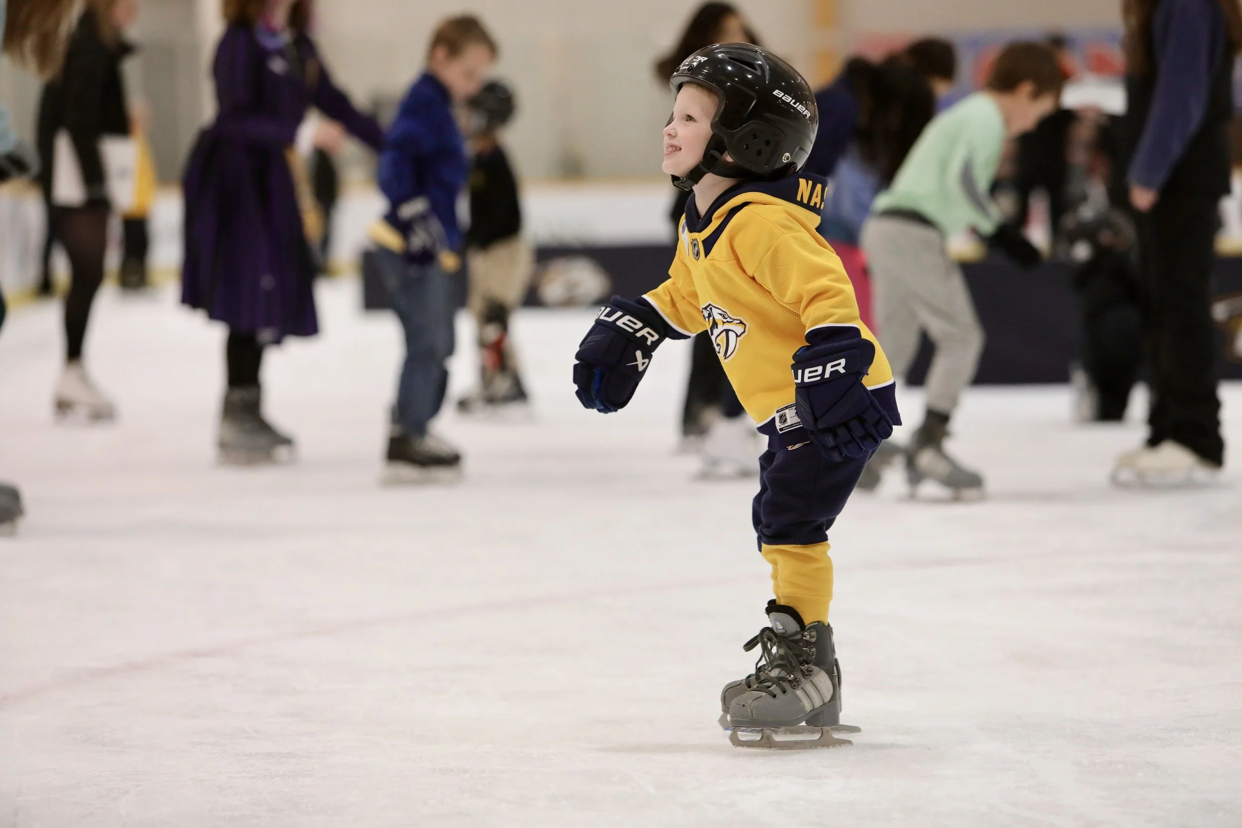 A young boy in a yellow hockey jersey and black helmet ice skating at an indoor rink, smiling and leaning forward slightly as he glides on his skates.