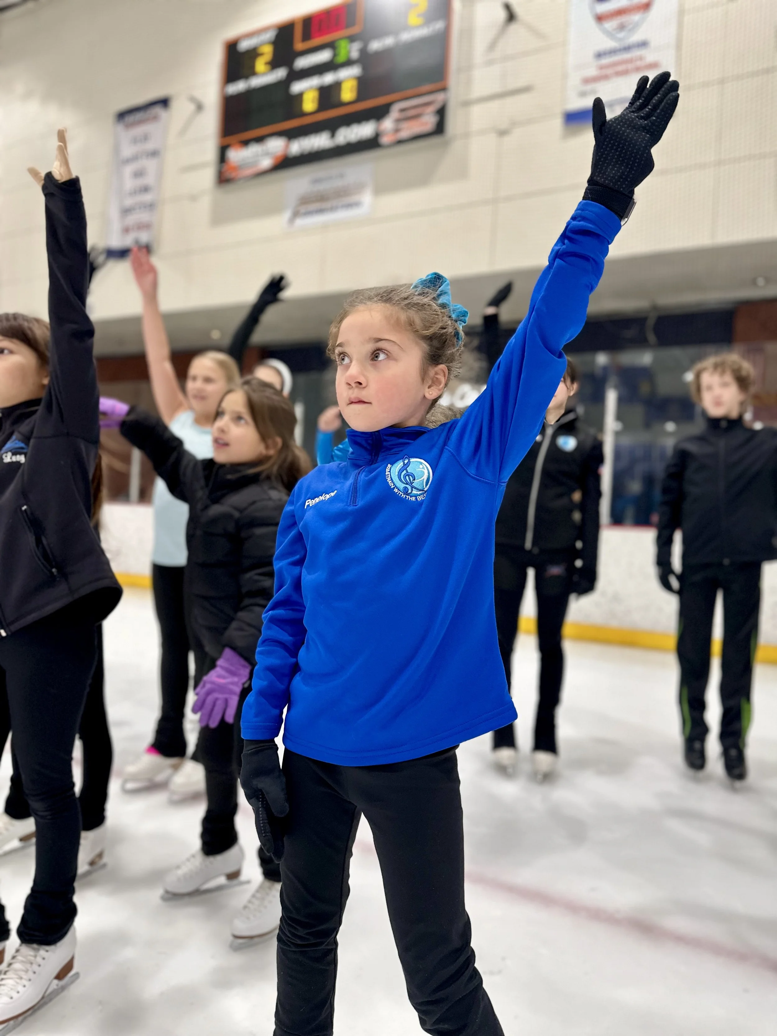 A young girl in a blue jacket with a blue scrunchie in her hair and black gloves, standing on ice rink with her arm raised during a skating class or practice, surrounded by other children also on ice.