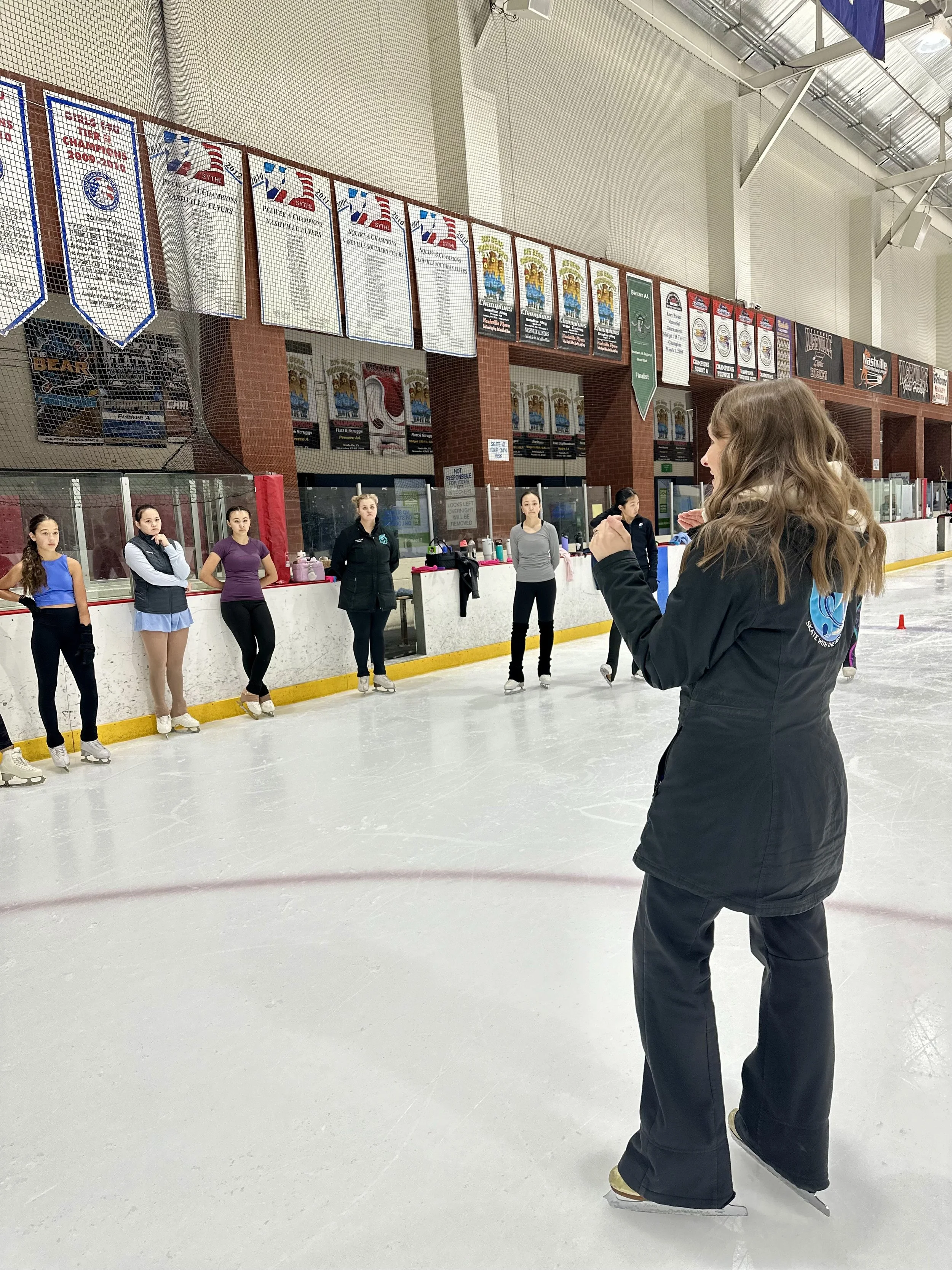 A group of young female ice skaters standing on the ice, listening to an instructor giving directions inside a roller skating rink, with banners hanging on the wall in the background.