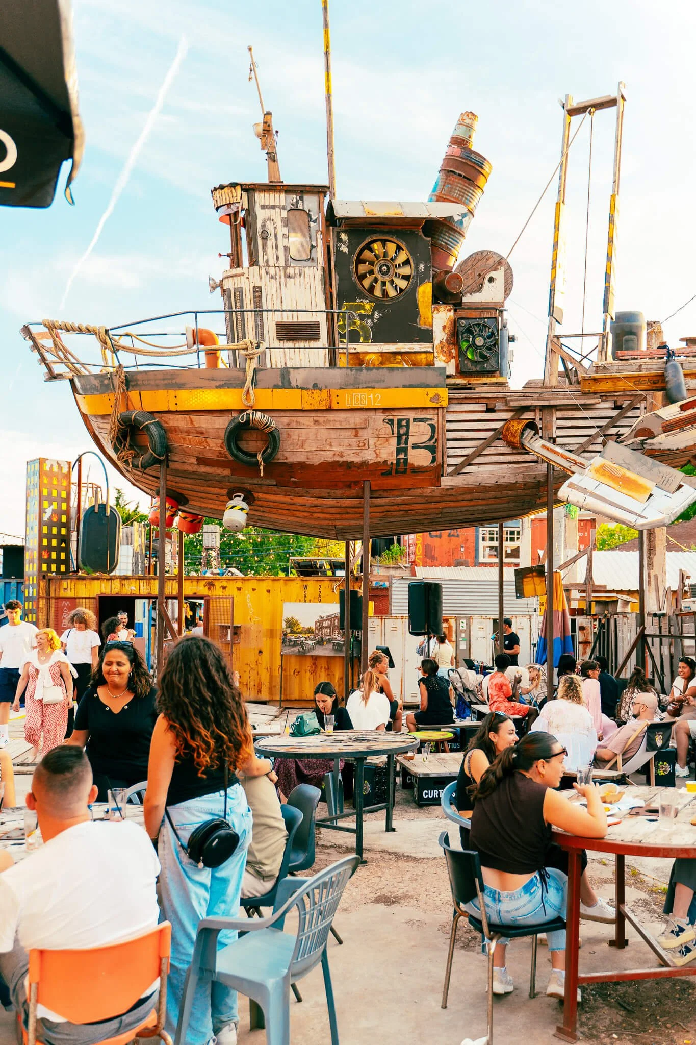 Un bâtiment en forme de bateau en bois flotté suspendu dans un espace extérieur où des personnes se détendent et discutent autour de tables.