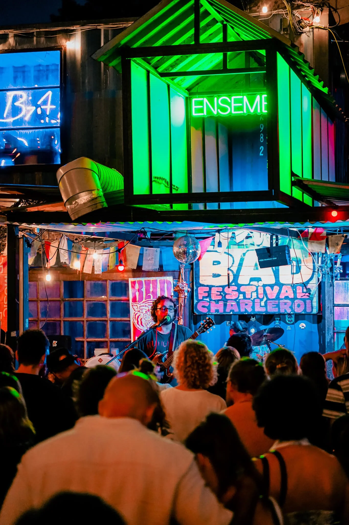 Une scène de concert en intérieur avec un groupe de musique jouant devant une foule. La scène est décorée de guirlandes et de signes lumineux, avec un panneau indiquant Festival Charlier.