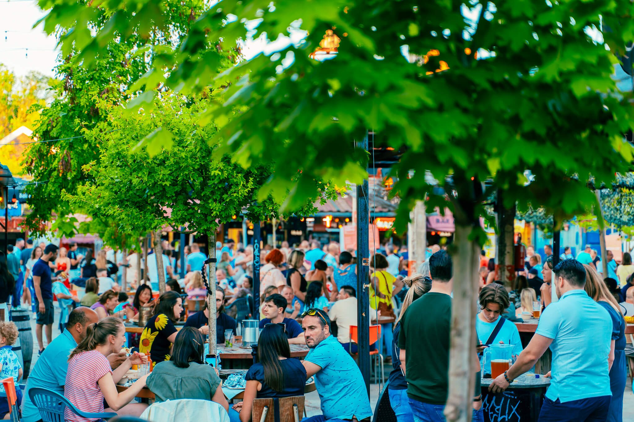 Une foule de personnes assises et debout dans une place animée en plein air, entourée d'arbres verts et de petits commerces. Certaines personnes mangent, d'autres discutent, créant une ambiance conviviale et festive.
