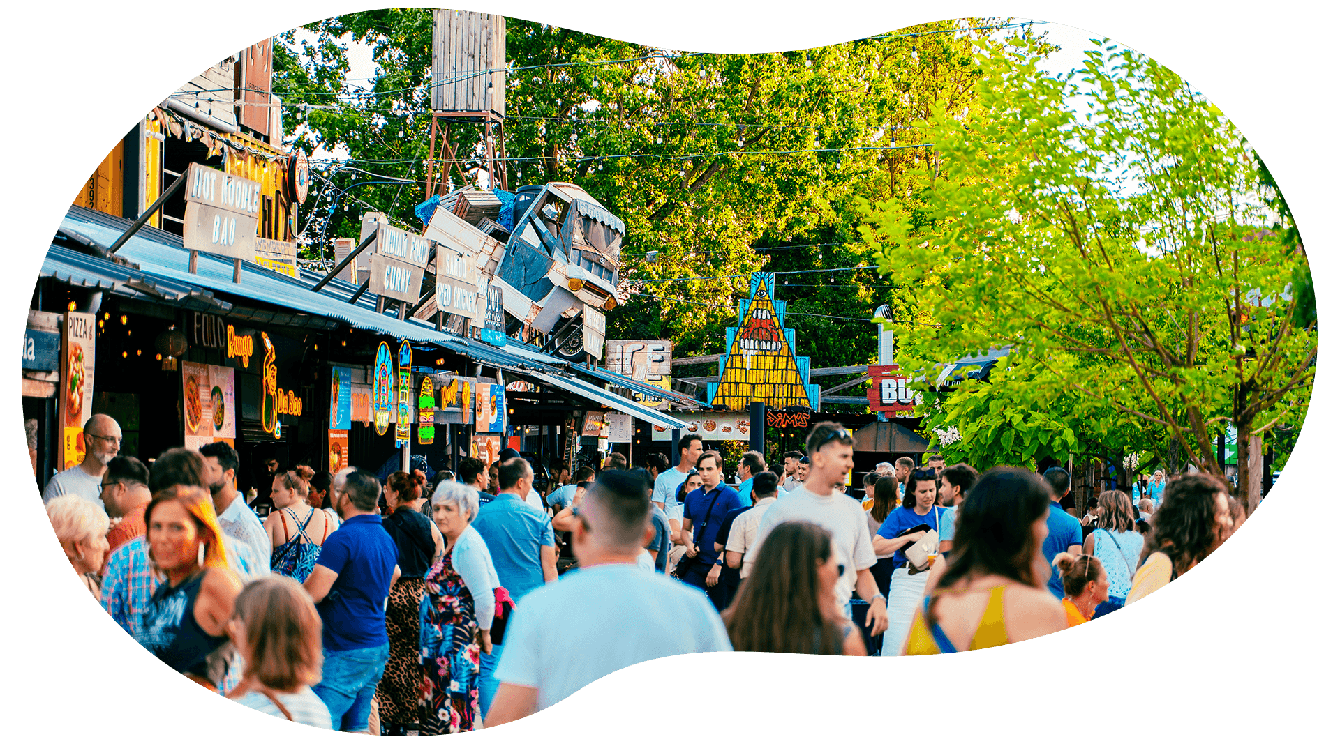 Foule de personnes dans une fête foraine ou marché en plein air avec manèges et stands, arbres verts en toile de fond, et décorations lumineuses.