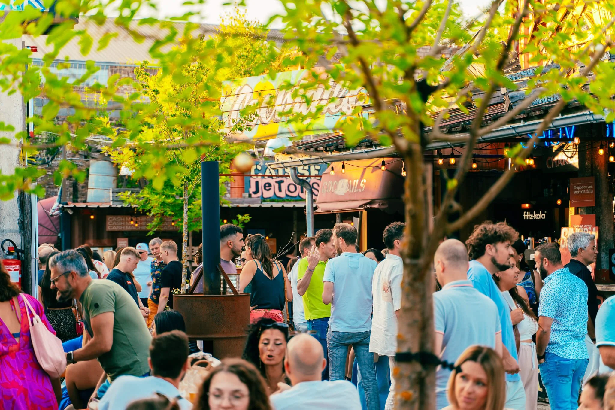 Une foule de personnes dans une rue animée lors d'un événement en plein air, avec des arbres en avant-plan et des enseignes lumineuses en arrière-plan.