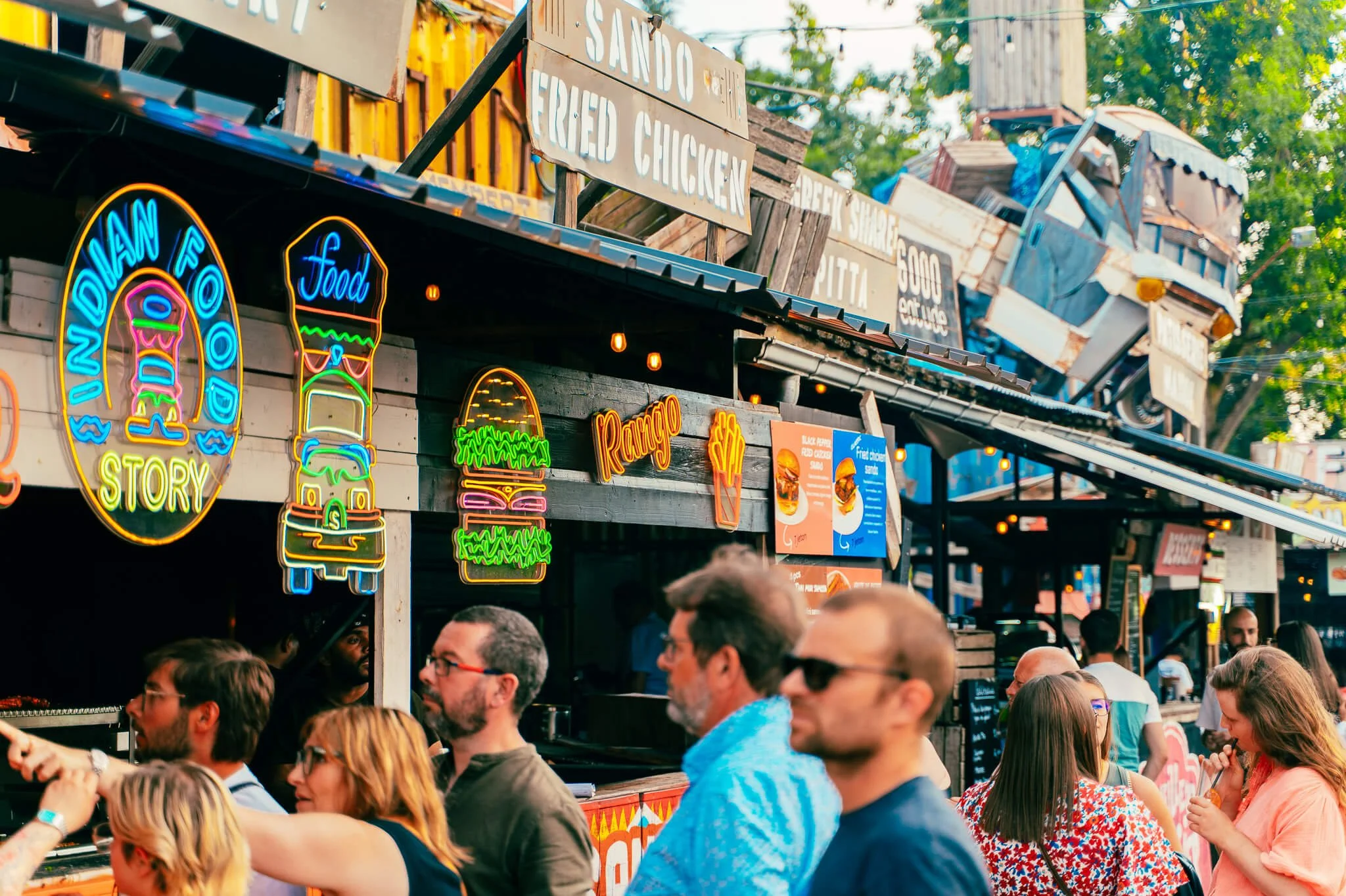 Stand de nourriture avec des panneaux en néon pour des hamburgers, poulet, steak et chili, avec des gens faisant la queue à l'extérieur, ambiance festive en plein air.