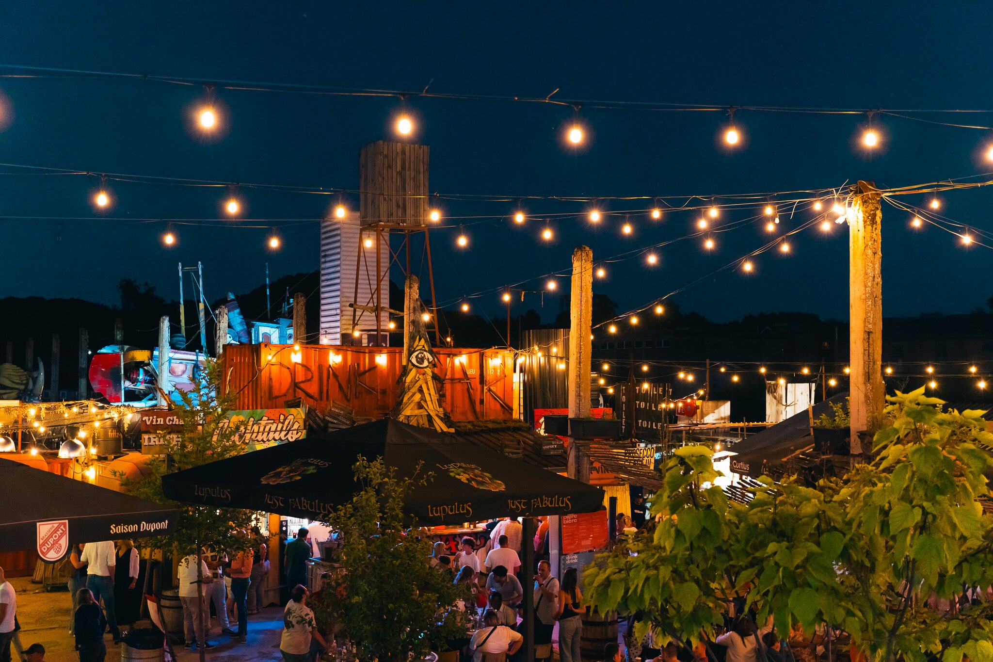 Une scène de fête extérieur la nuit avec des guirlandes lumineuses, de nombreuses personnes, des tables, des parasols et des bâtiments en arrière-plan.