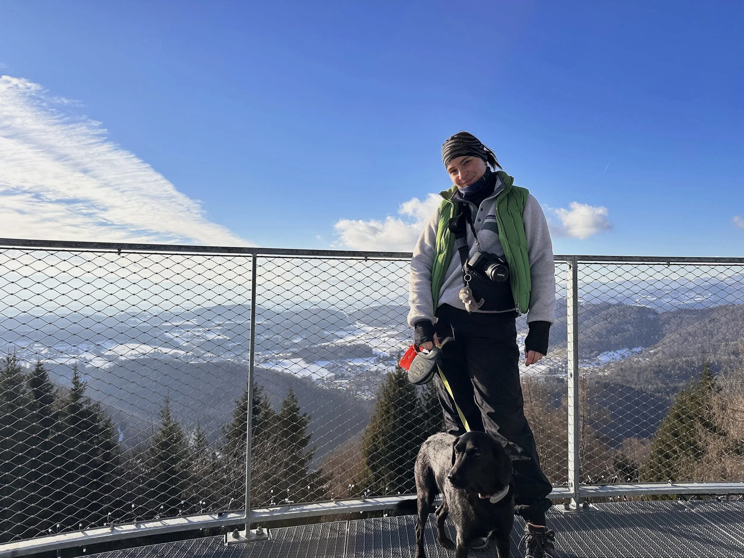 Ein Mann mit schwarzem Hund und einer Dodger-Tasche steht auf einer Aussichtsplattform mit einem Drahtzaun, im Hintergrund sind Wolken und Berge sichtbar. Es ist eine klare, sonnige Wintertag.
