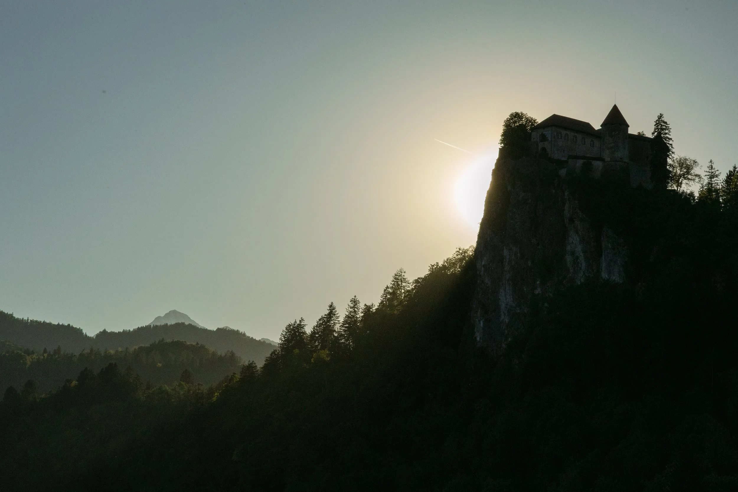 Burg auf einem Hügel bei Sonnenuntergang, umgeben von Bäumen und Bergen