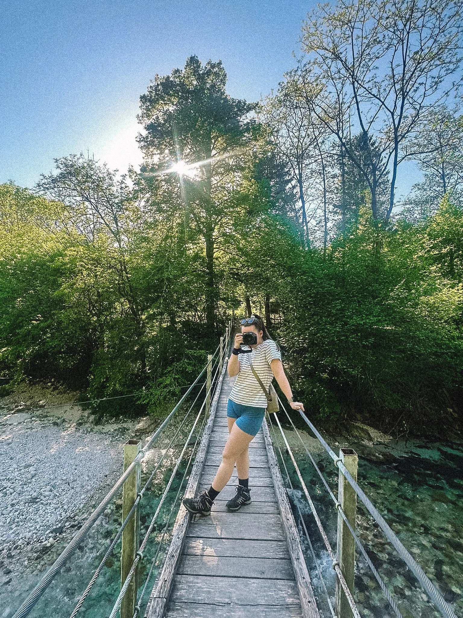 Junge Frau mit Sonnenbrille, gestreiftem T-Shirt, Jeansshorts und Wanderschuhen fotografiert sich auf einer Hängebrücke in einem Wald. Die Sonne scheint durch die Bäume.
