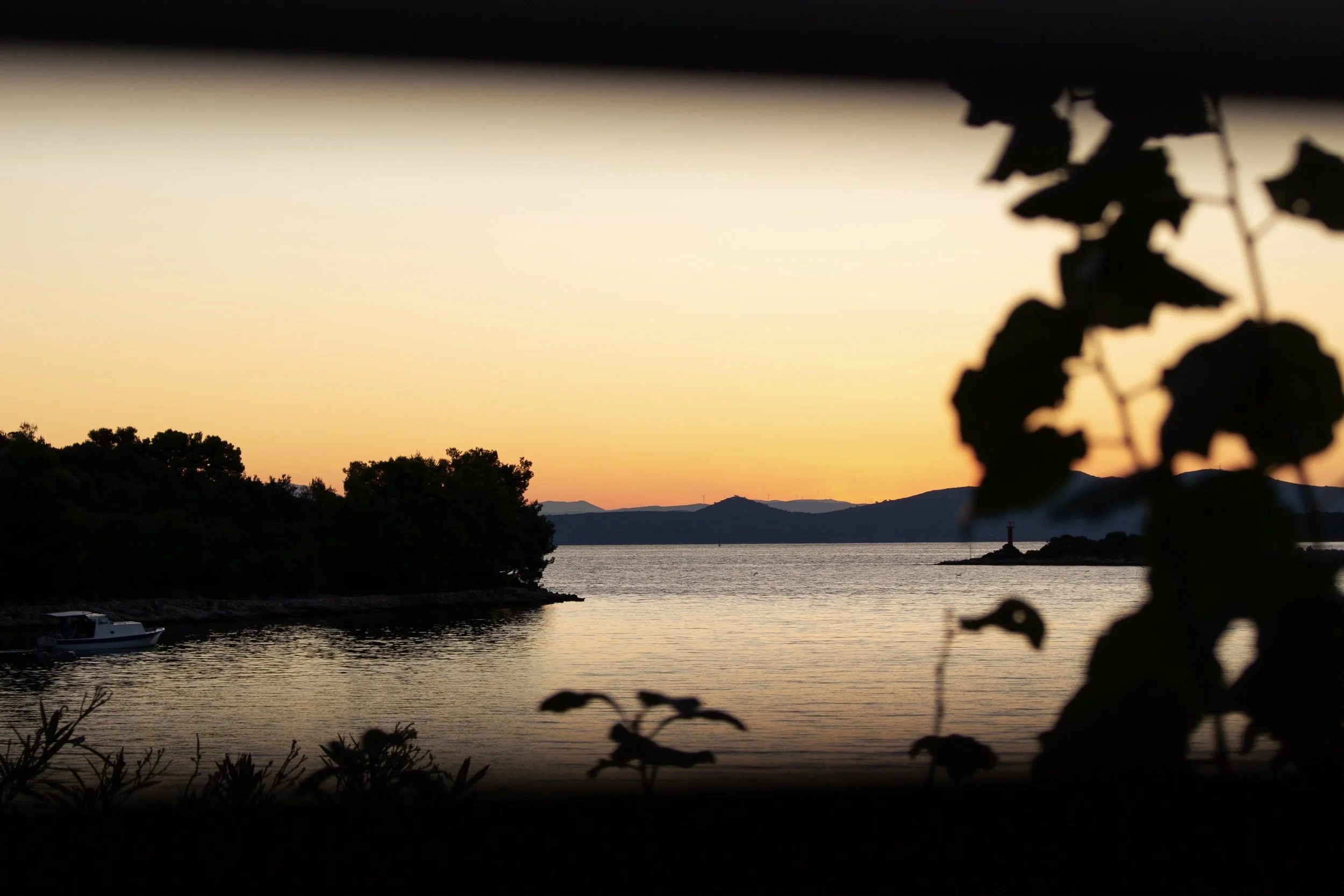 Sonnenuntergang über einem ruhigen Wasser mit einem Leuchtturm auf einer kleinen Insel, silhouettierte Bäume im Vordergrund.