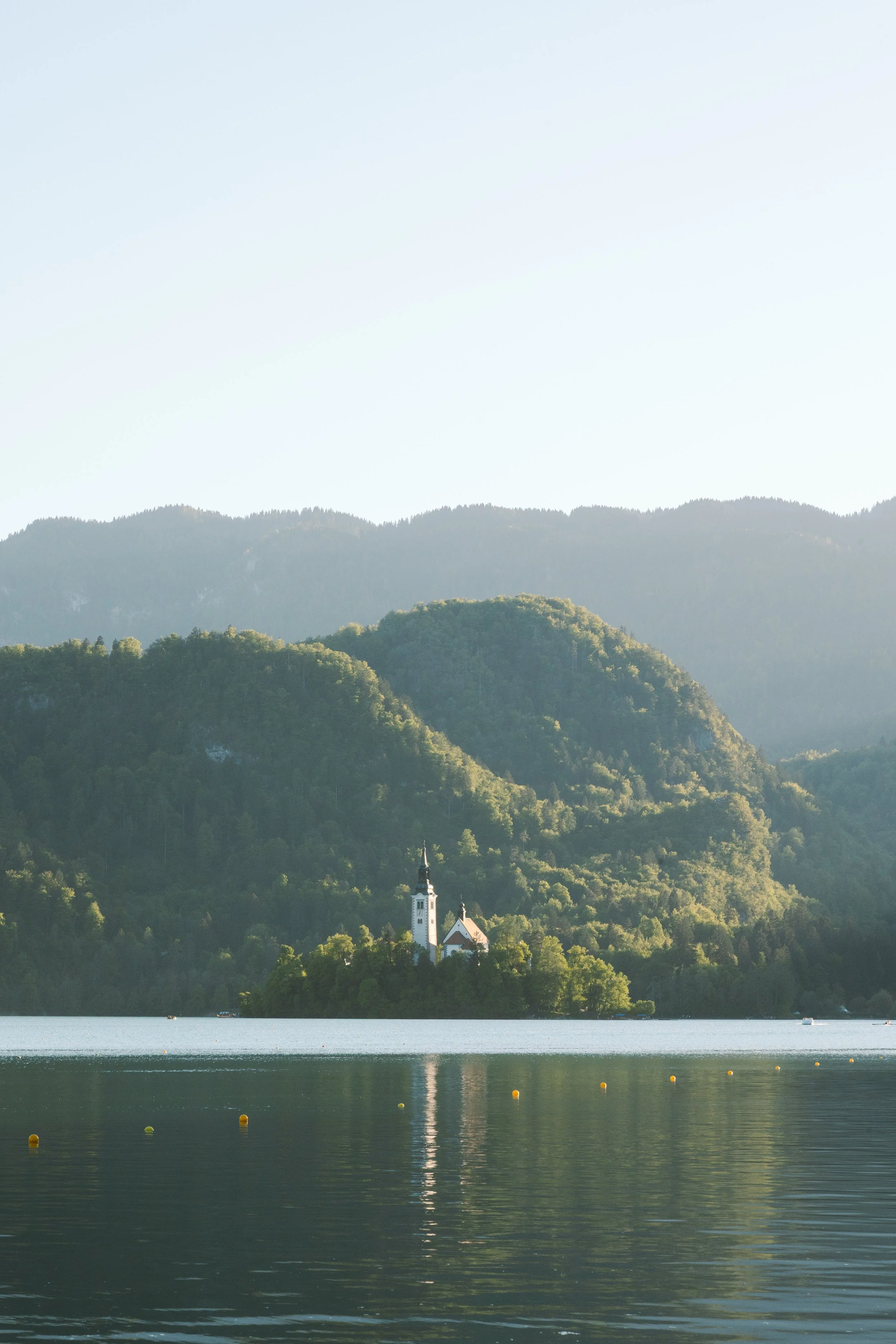 Ein See mit einem Ufer im Vordergrund, im Hintergrund Hügel und Berge mit einem kleinen weißen Kirche auf einem Hügel, umgeben von Bäumen, unter einem klaren Himmel.