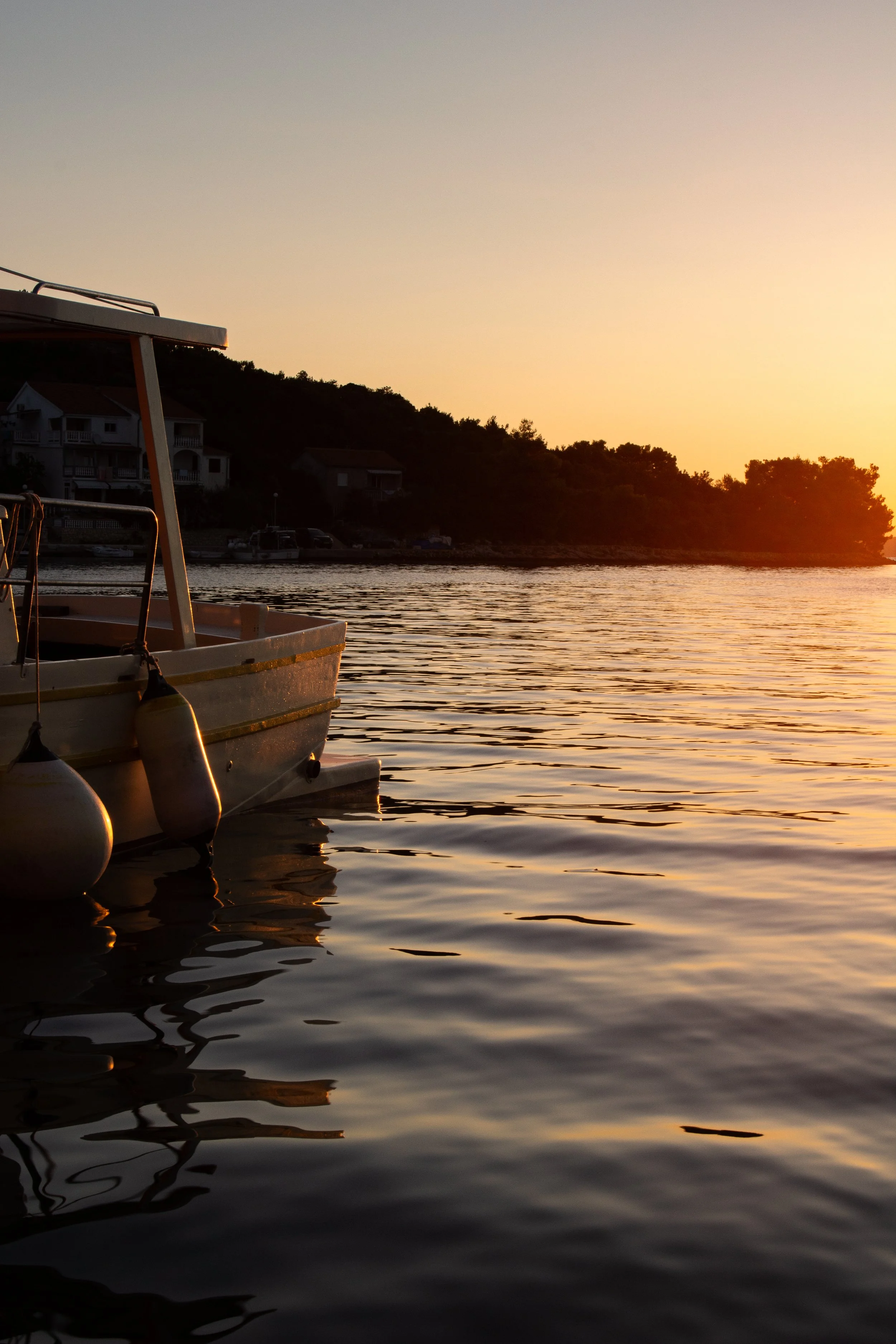 Ein Boot liegt auf ruhigem Wasser bei Sonnenuntergang, im Hintergrund sind Hügel mit Häusern und Bäumen sichtbar.