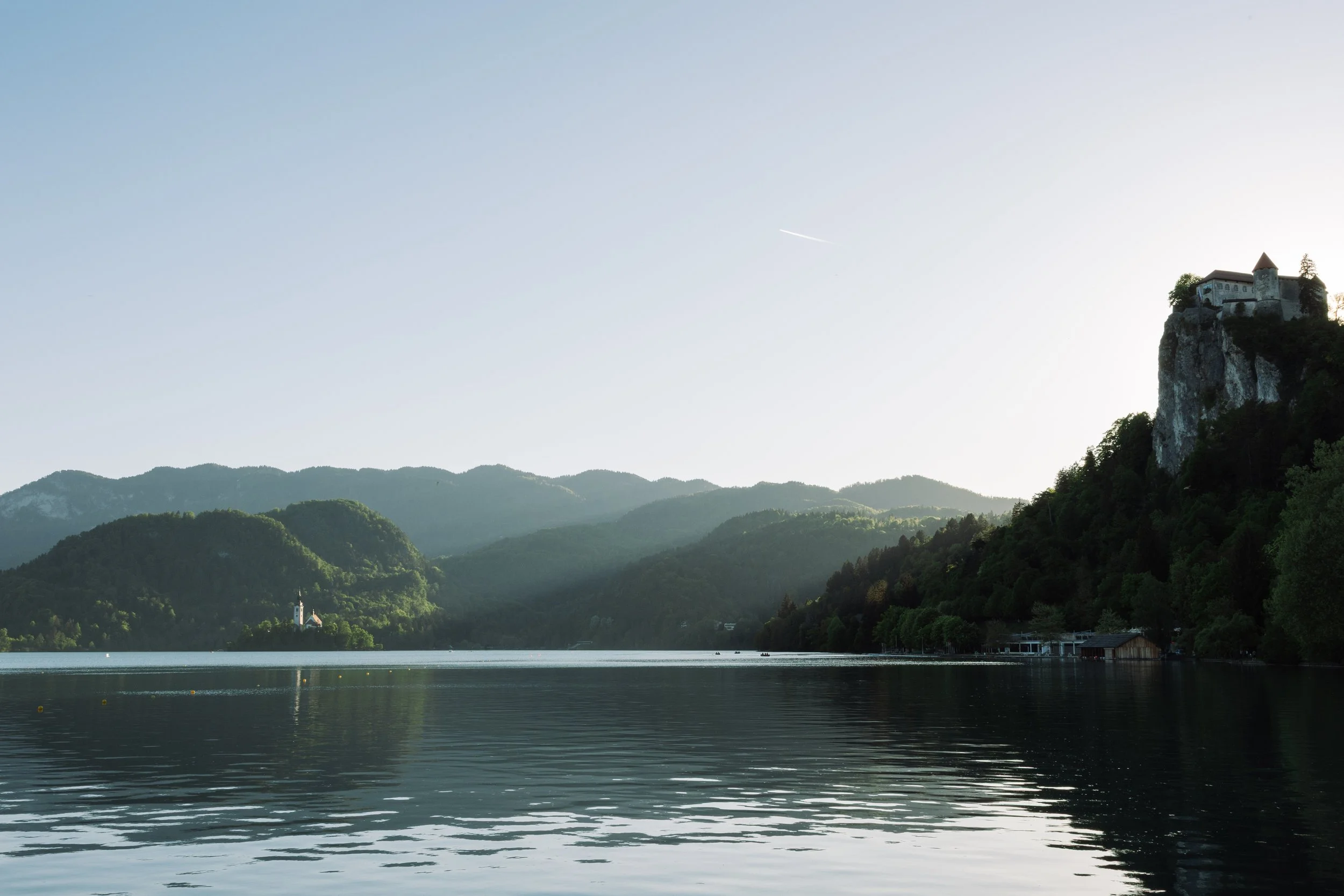 Blick auf einen See mit Bergen im Hintergrund und eine kleine Burg auf einem Felsen rechts im Bild, bei Sonnenuntergang