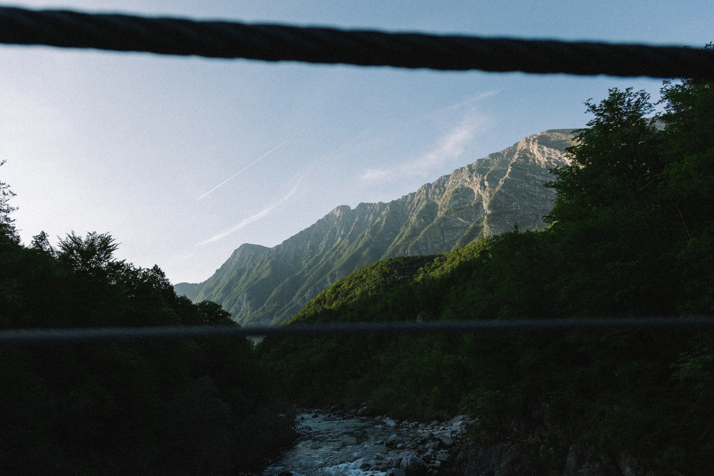 Berglandschaft mit Fluss im Vordergrund, Bäume und hohe Berge im Hintergrund, blauer Himmel.