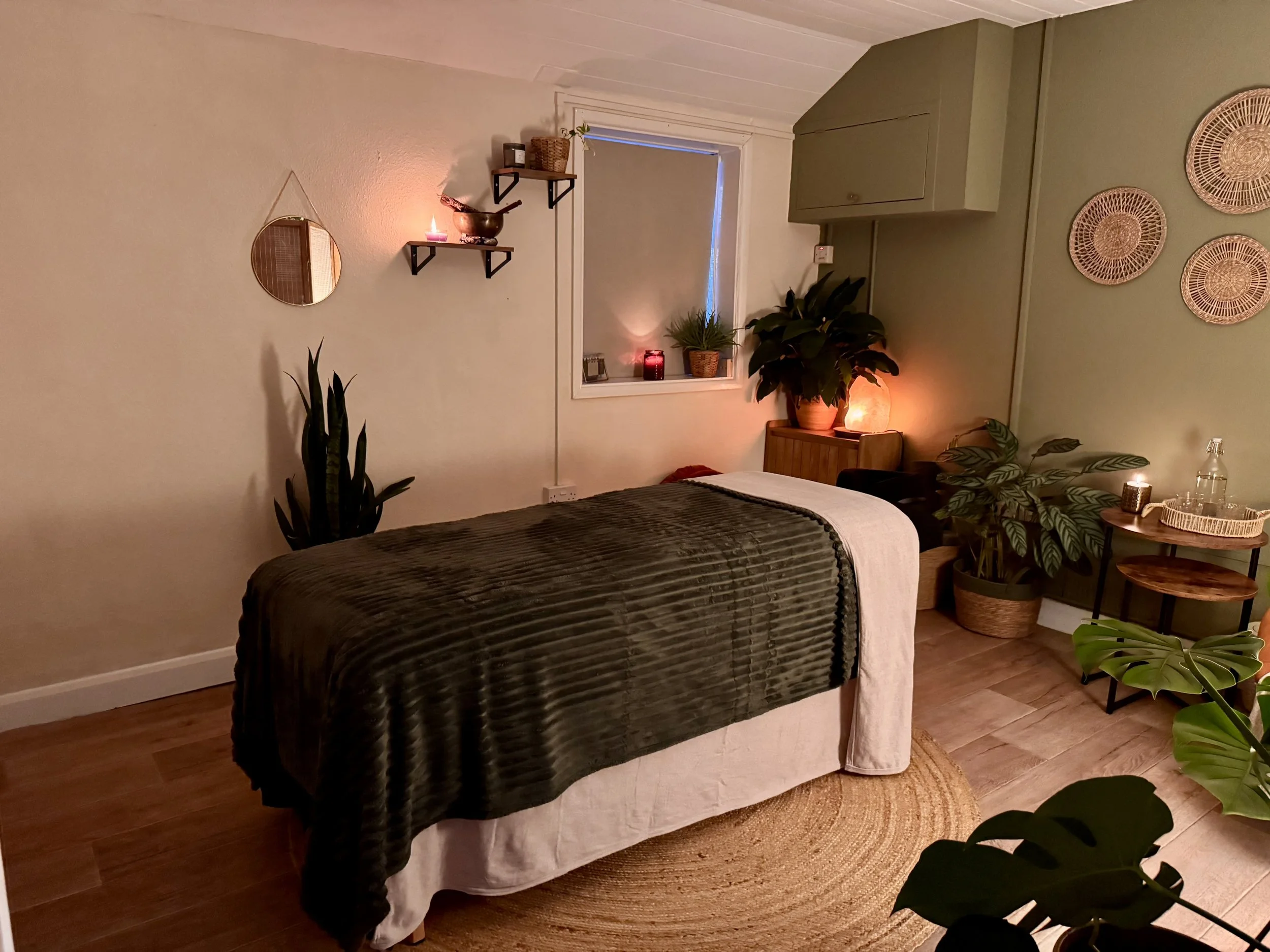 A cozy room with a massage table covered with a dark green blanket, surrounded by plants, candles, and decorative wall art, illuminated with warm lighting.