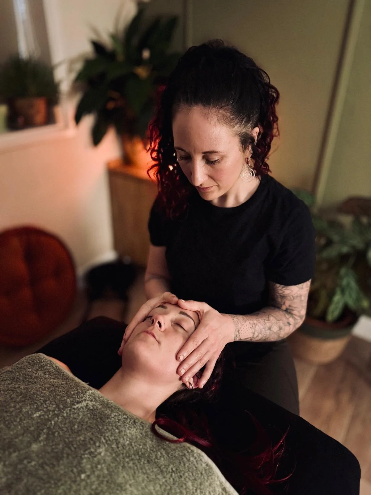 A woman receiving a facial massage from a masseuse in a cozy room with plants and warm lighting.