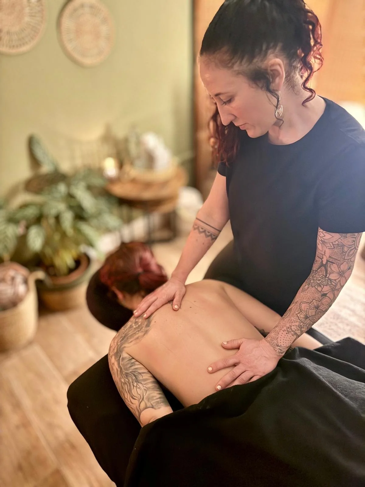 A woman giving a massage to a person lying face down on a massage table in a cozy room with potted plants and decorative plates on the wall.