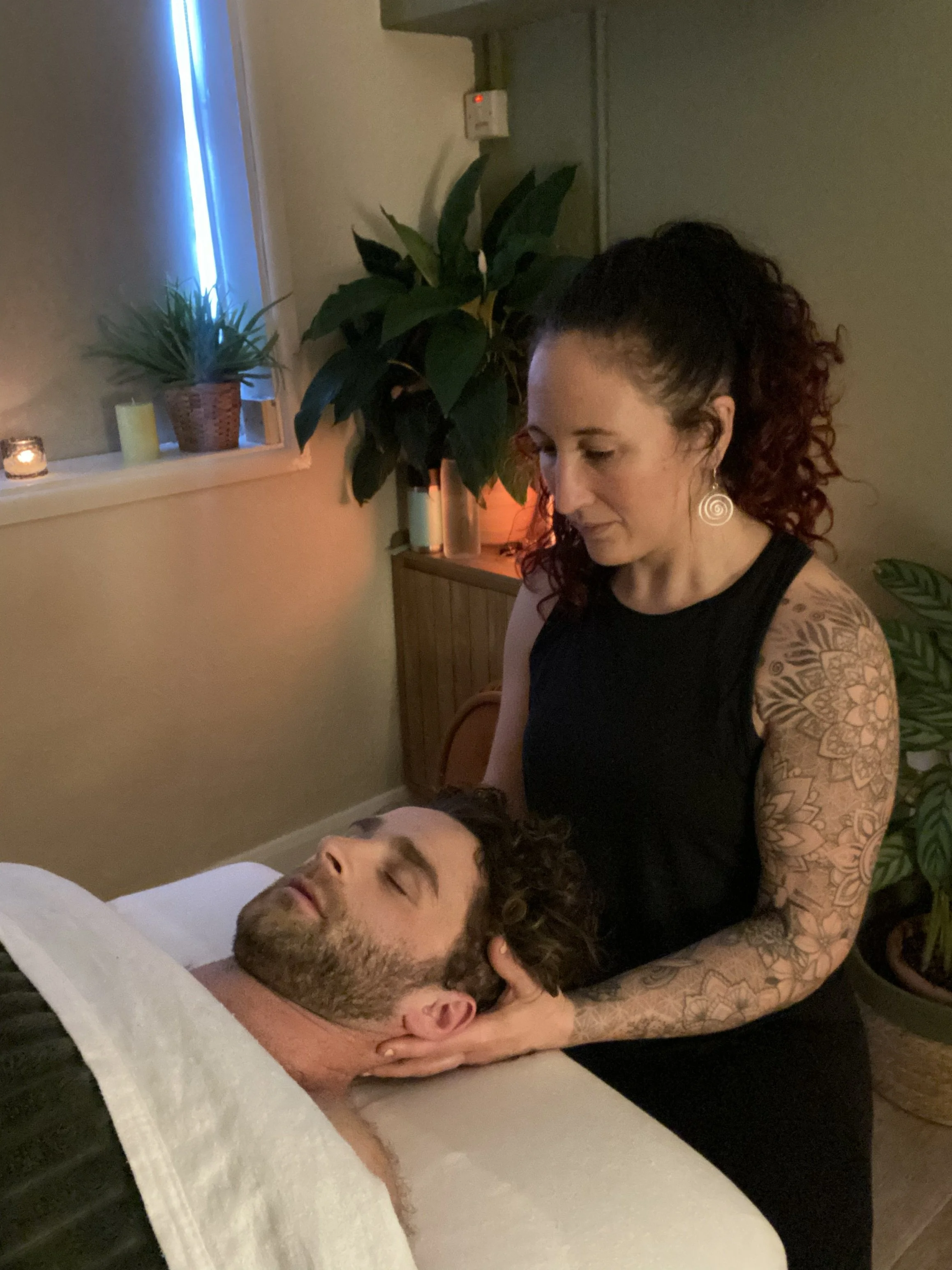 A woman with curly hair and floral tattoos on her arm gives a face massage to a man lying on a massage table in a cozy, softly lit room with plants and candles.