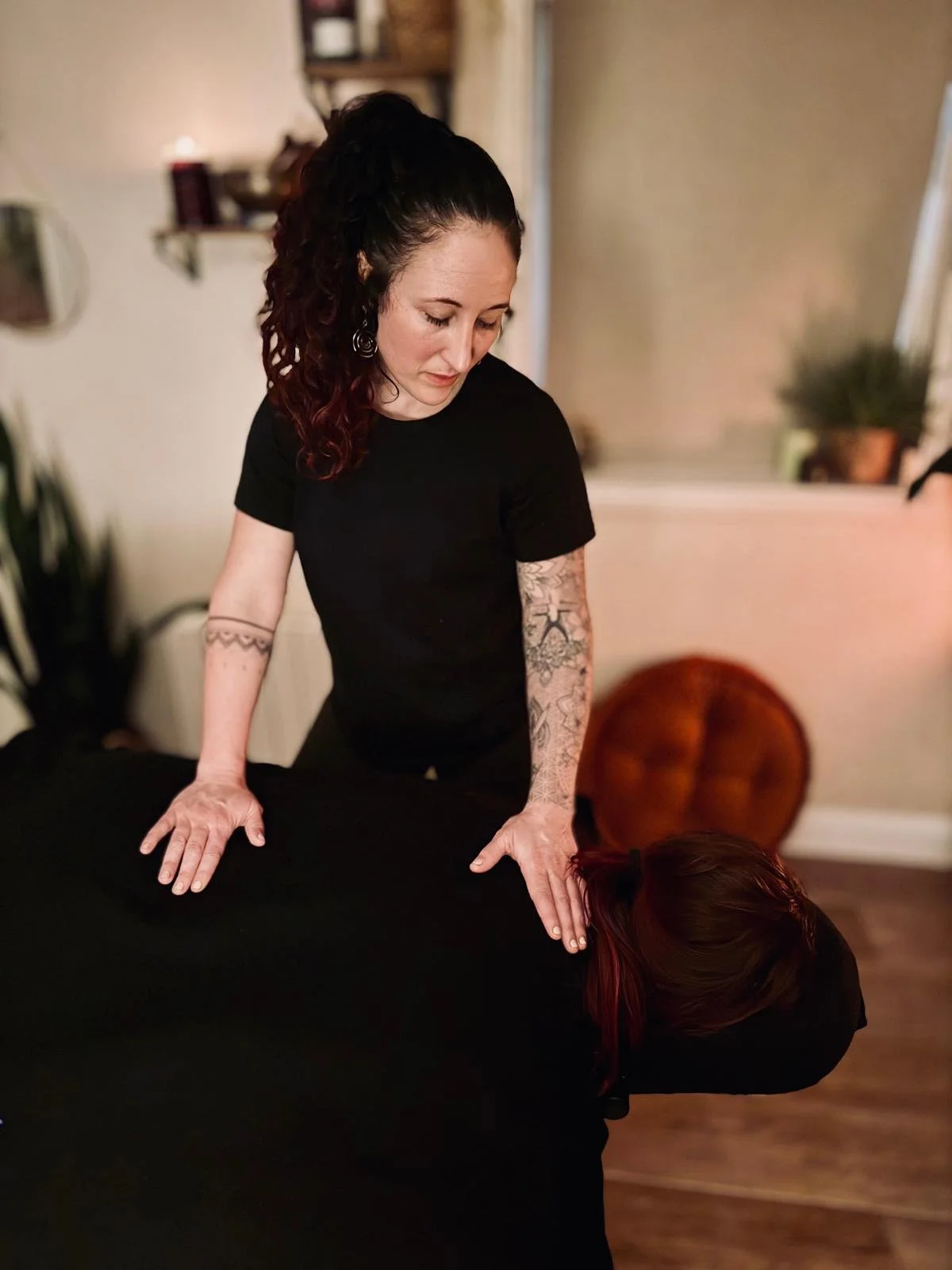 A woman gives a massage to a person lying face down on a massage table in a cozy room, with plants and decorations in the background.