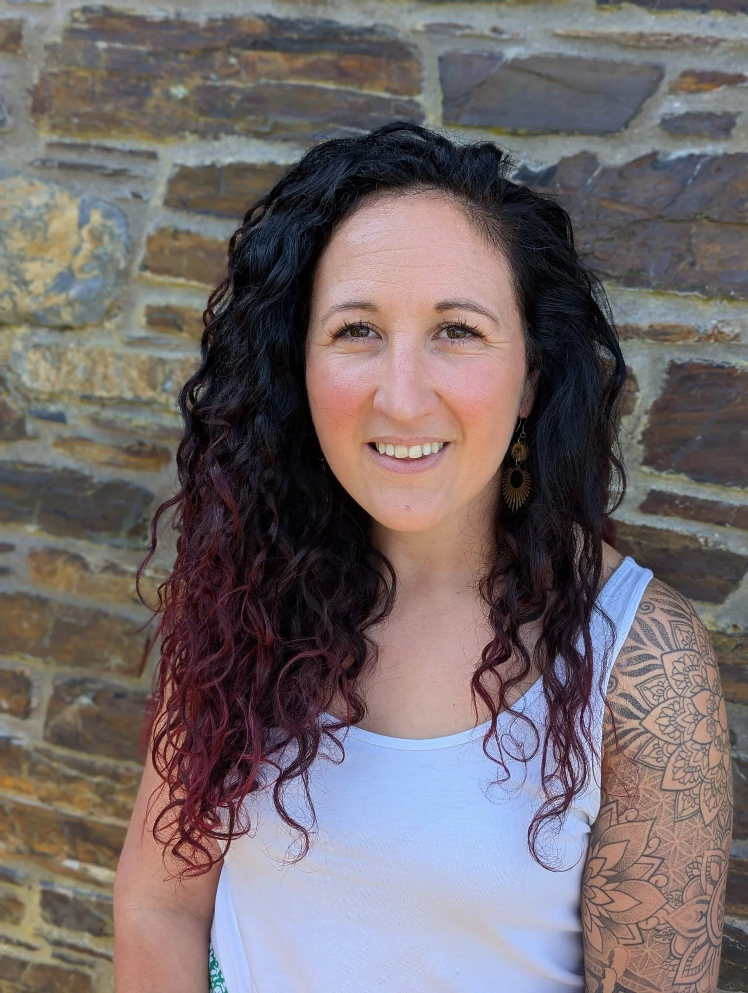 A woman with curly black hair and pink tips, wearing a white sleeveless top, earrings, and a tattoo sleeve on her right arm, smiling in front of a stone wall.