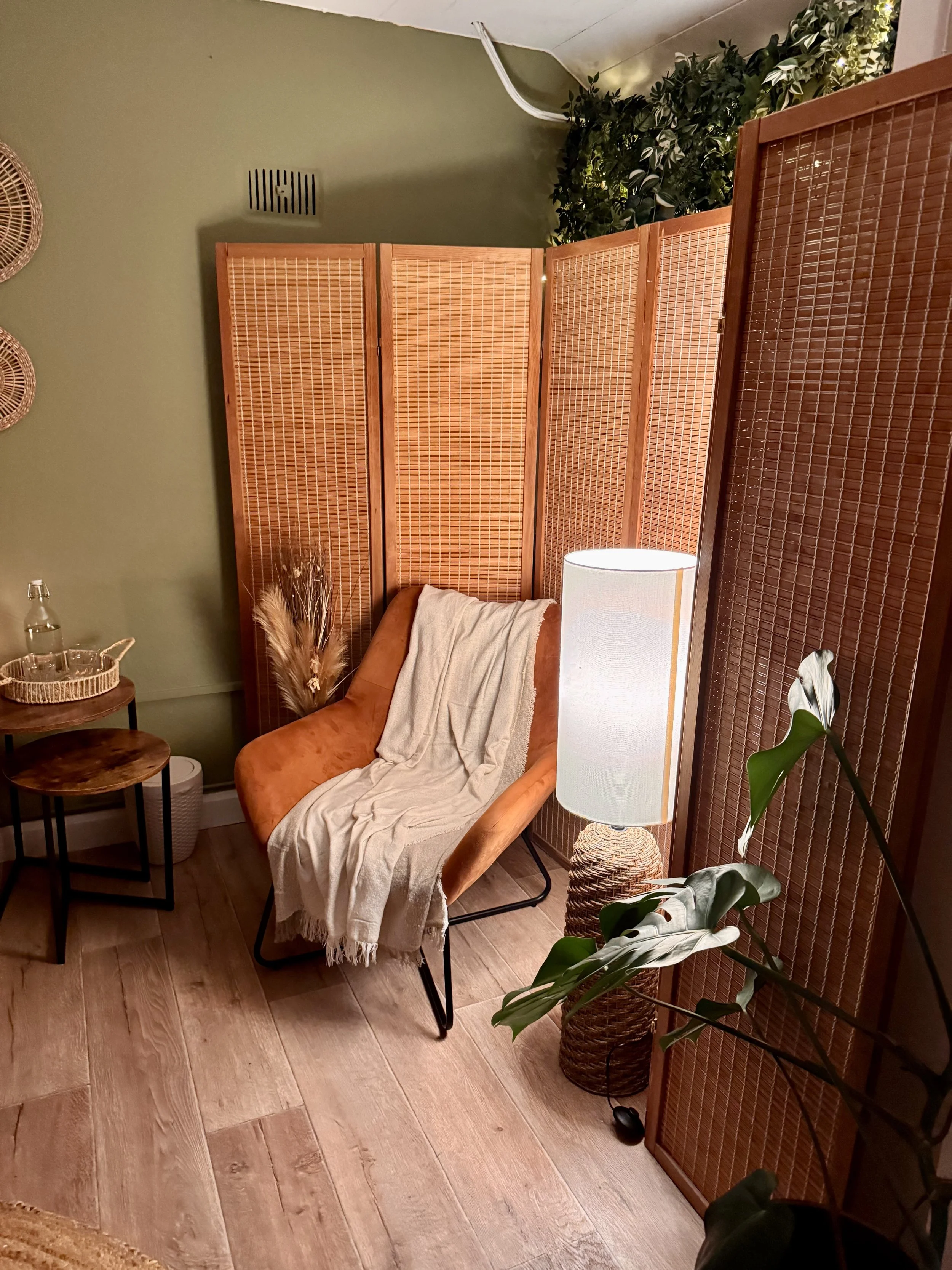 A cozy corner with a tan armchair covered with a beige throw blanket, a small wooden side table with a basket, a table lamp on a wicker base, a large plant with broad leaves, and a wooden room divider with woven panels, against a green wall.